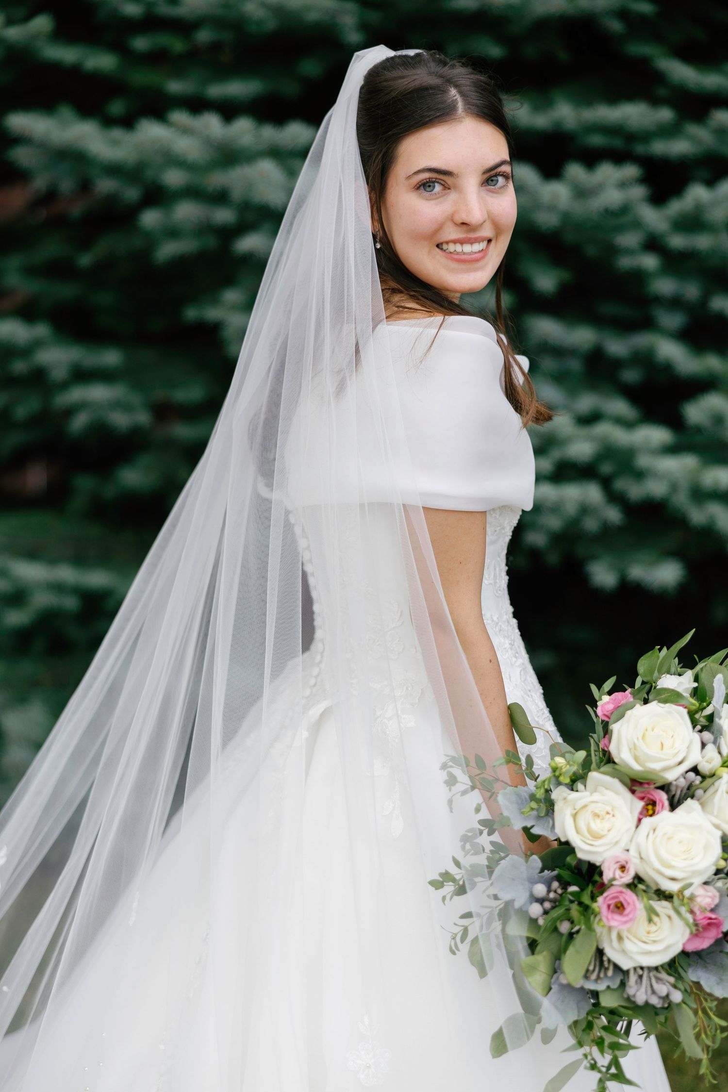 Smiling bride in white off-shoulder gown and veil holds floral bouquet outdoors near evergreen trees.