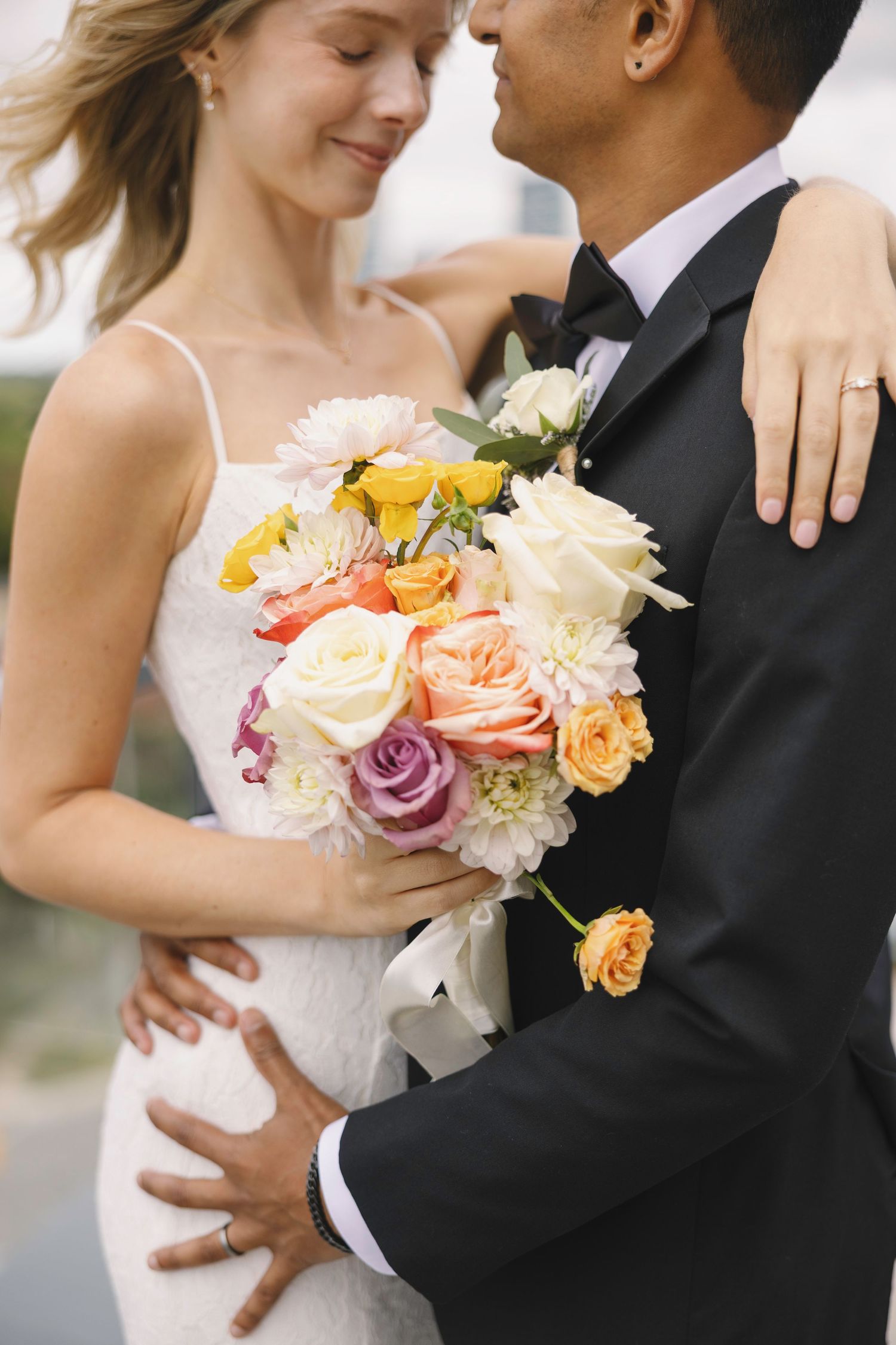 A bride and groom embrace closely, with the bride holding a colorful bouquet of roses and dahlias on their wedding day.