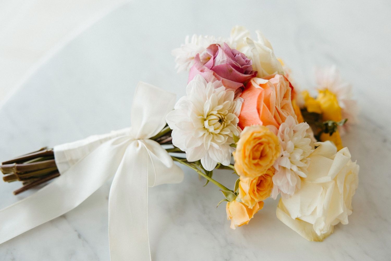 Elegant wedding bouquet with pink, orange, and white flowers tied with a white ribbon on marble.