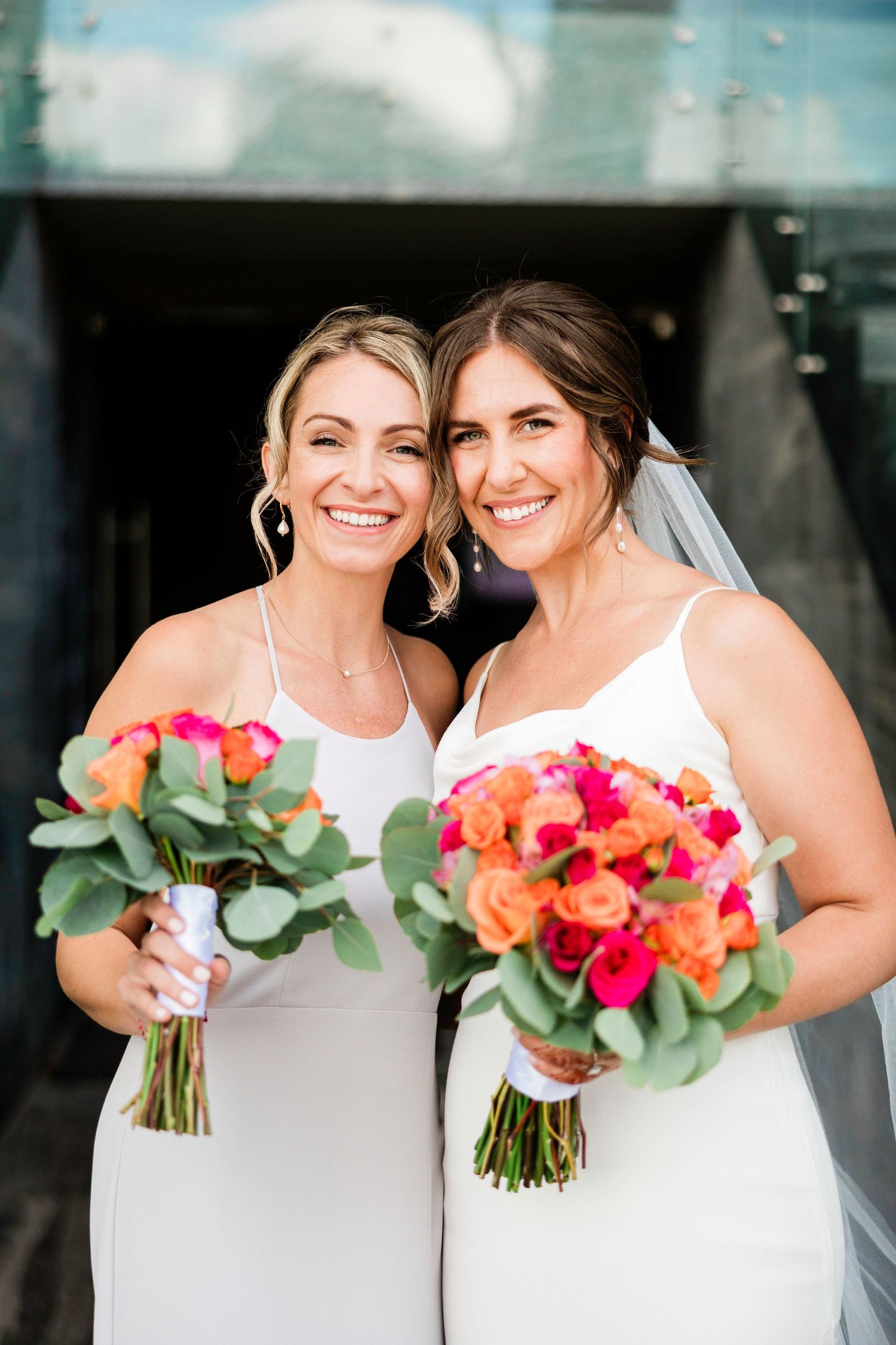 Two people in white dresses hold vibrant coral and pink wedding bouquets while smiling together.