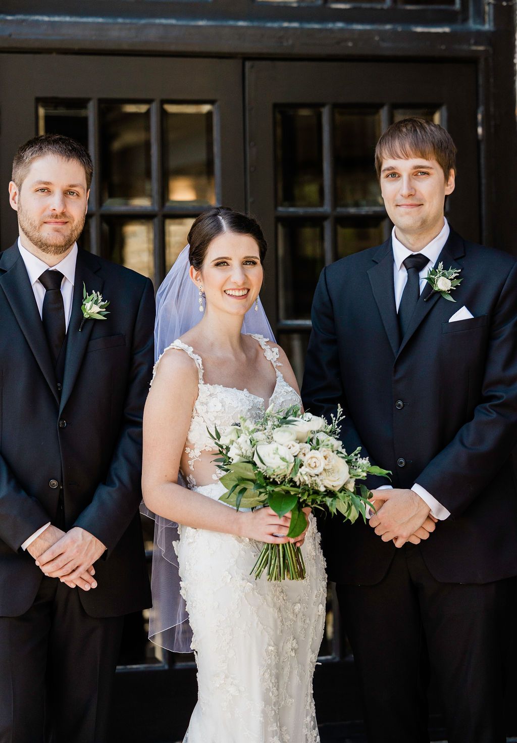 Wedding party in formal attire with black suits and white dress poses together outdoors.