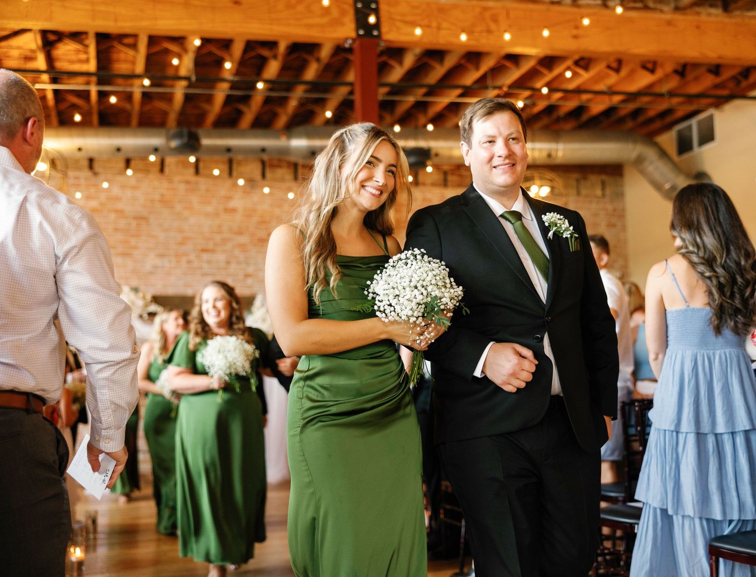 Wedding party members dance together in a rustic venue with exposed brick walls and string lights overhead.