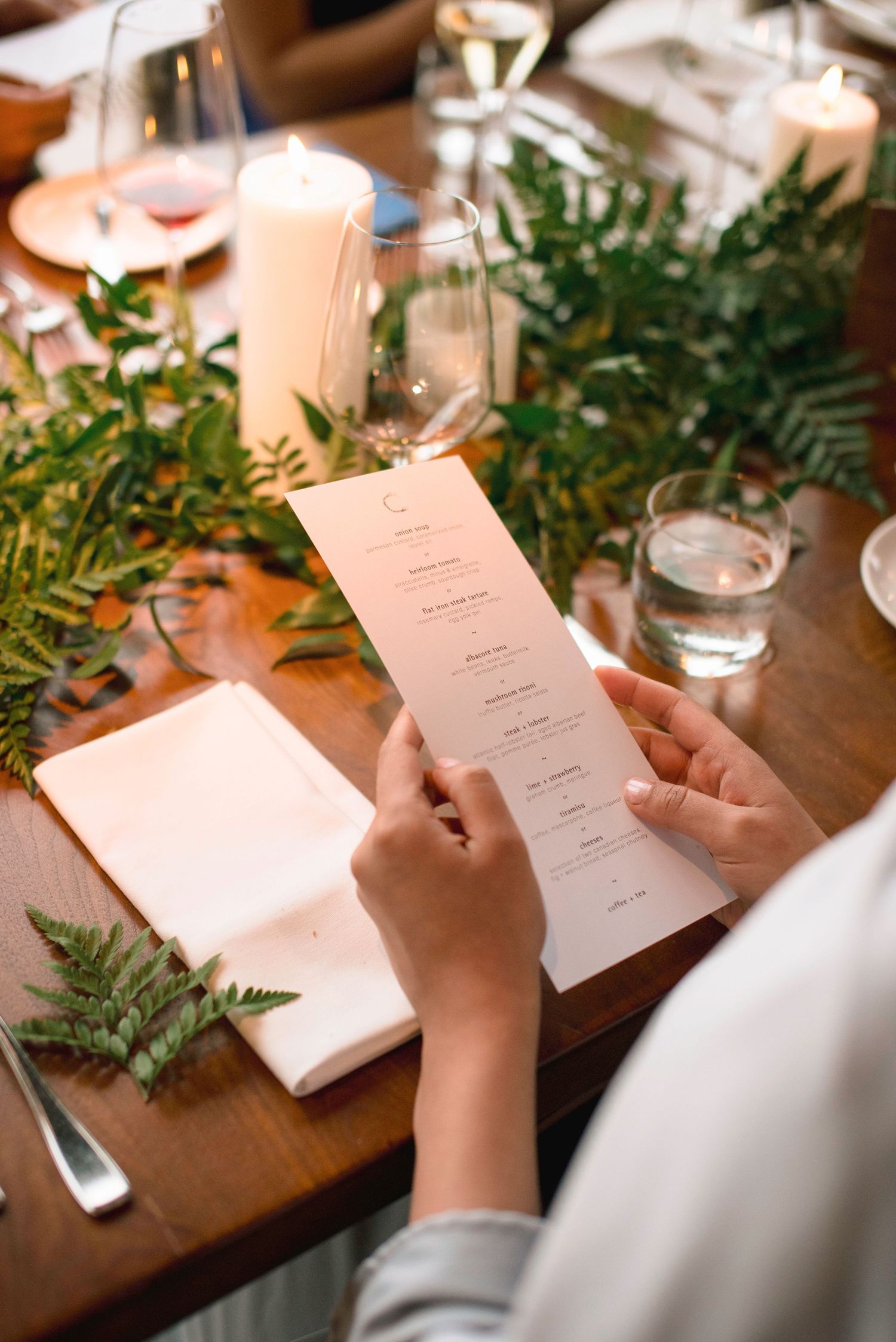 Hands holding wedding menu cards with green foliage decorations on wooden table.