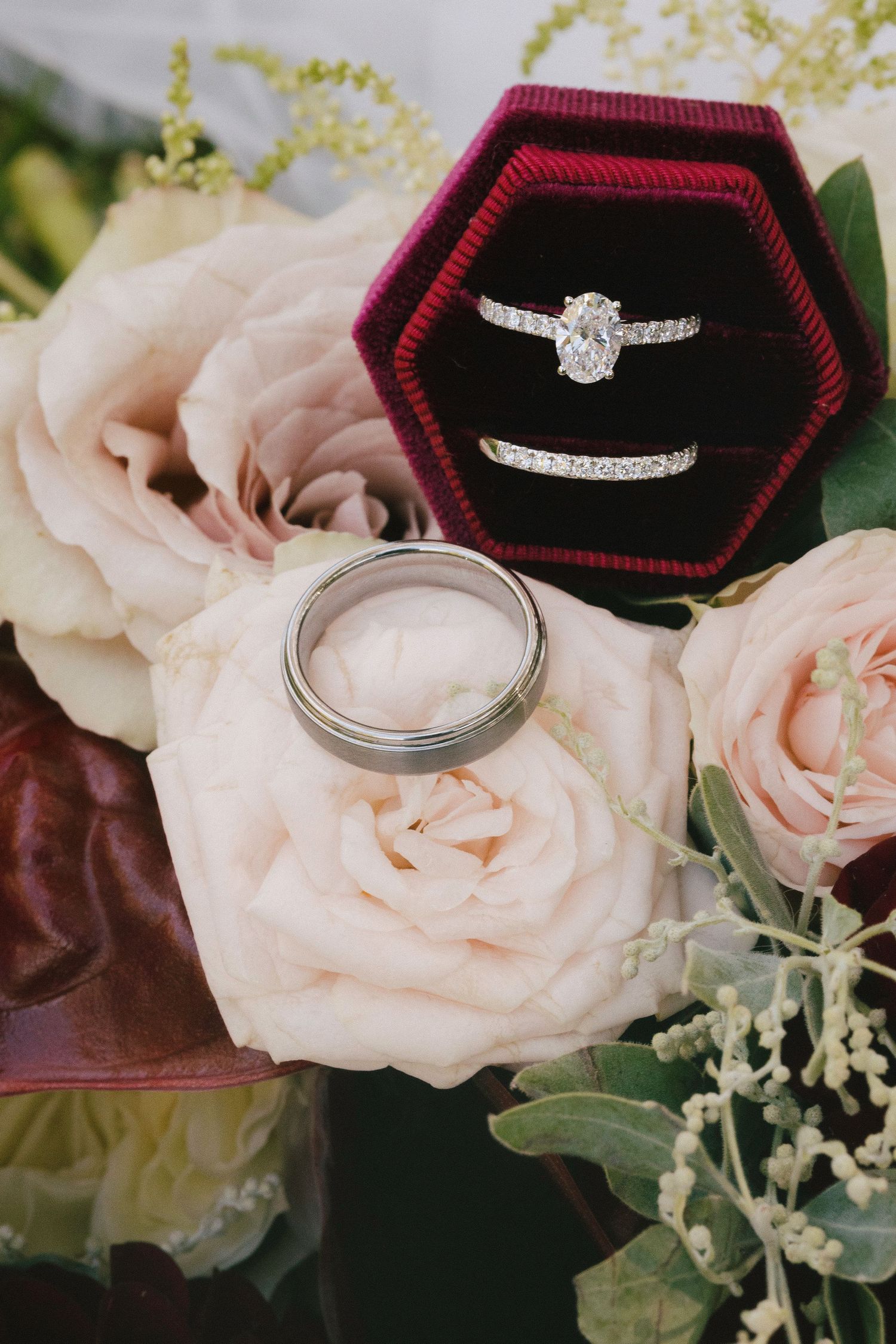 Wedding rings displayed on blush roses, featuring diamond engagement ring in red velvet box with matching band and silver groom's ring.