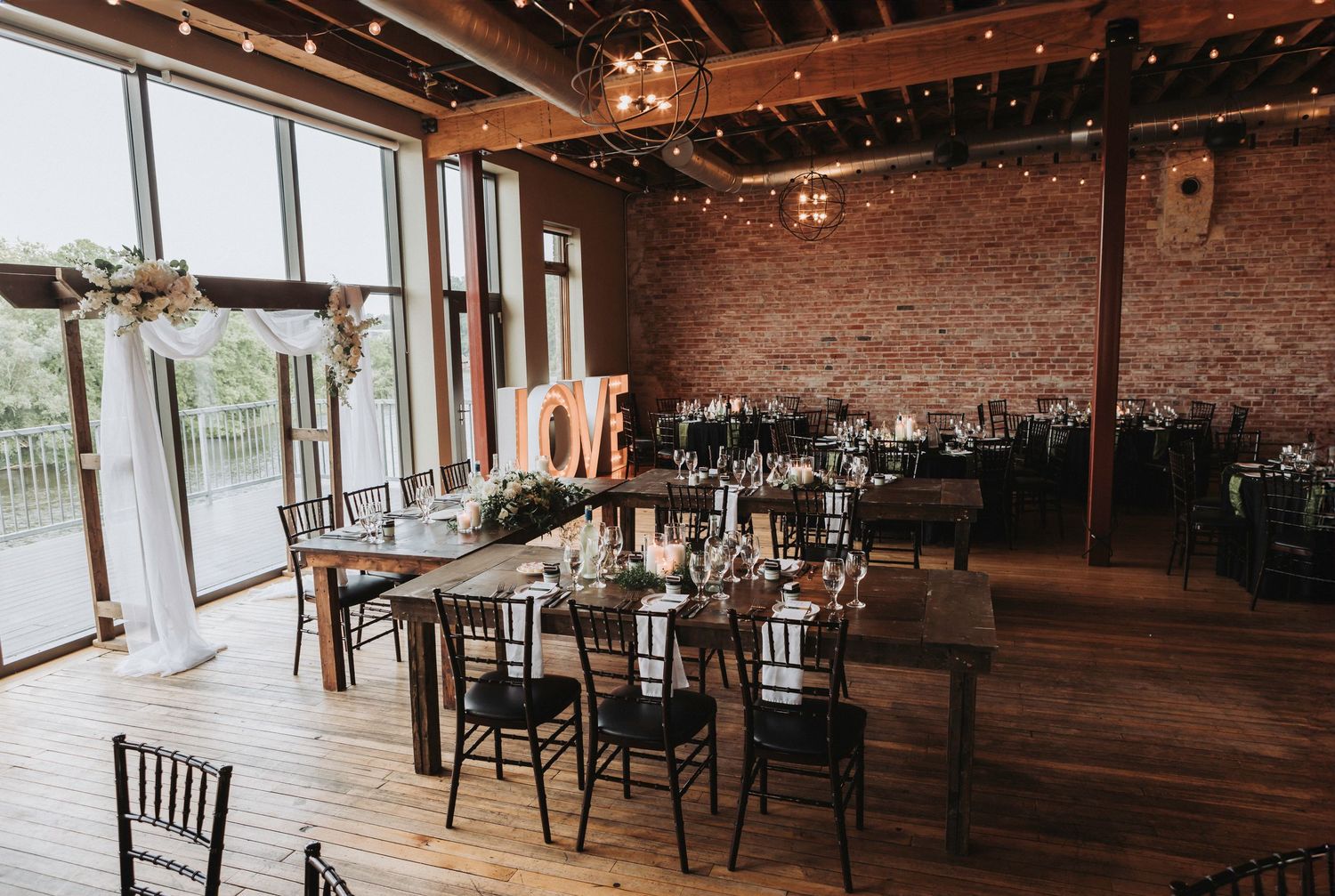 Rustic brick wall venue with wooden beams, string lights, and dark Chiavari chairs set up for an elegant event space.