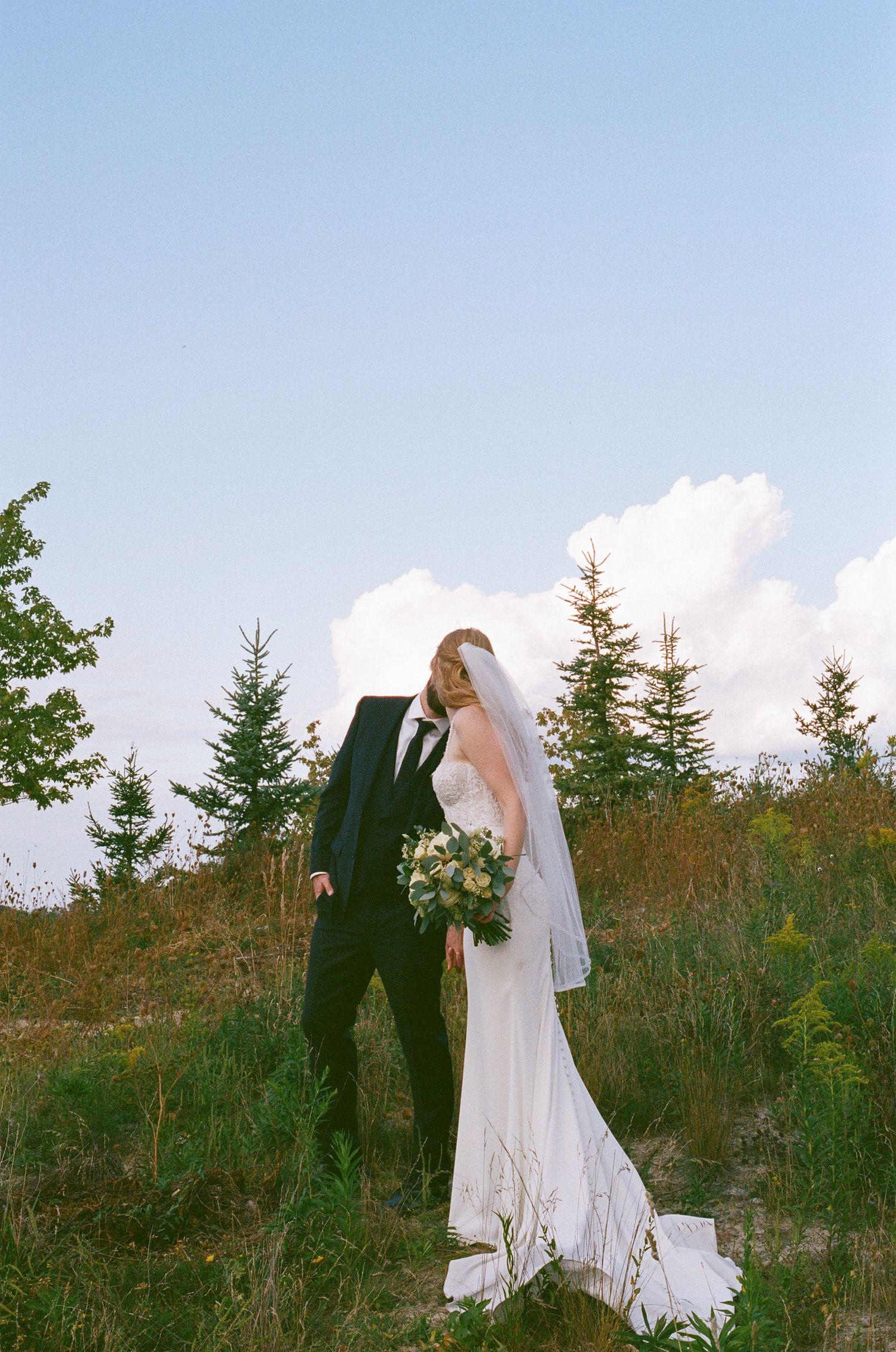 A wedding couple shares intimate moments on a hillside with evergreen trees in the background during their outdoor ceremony.