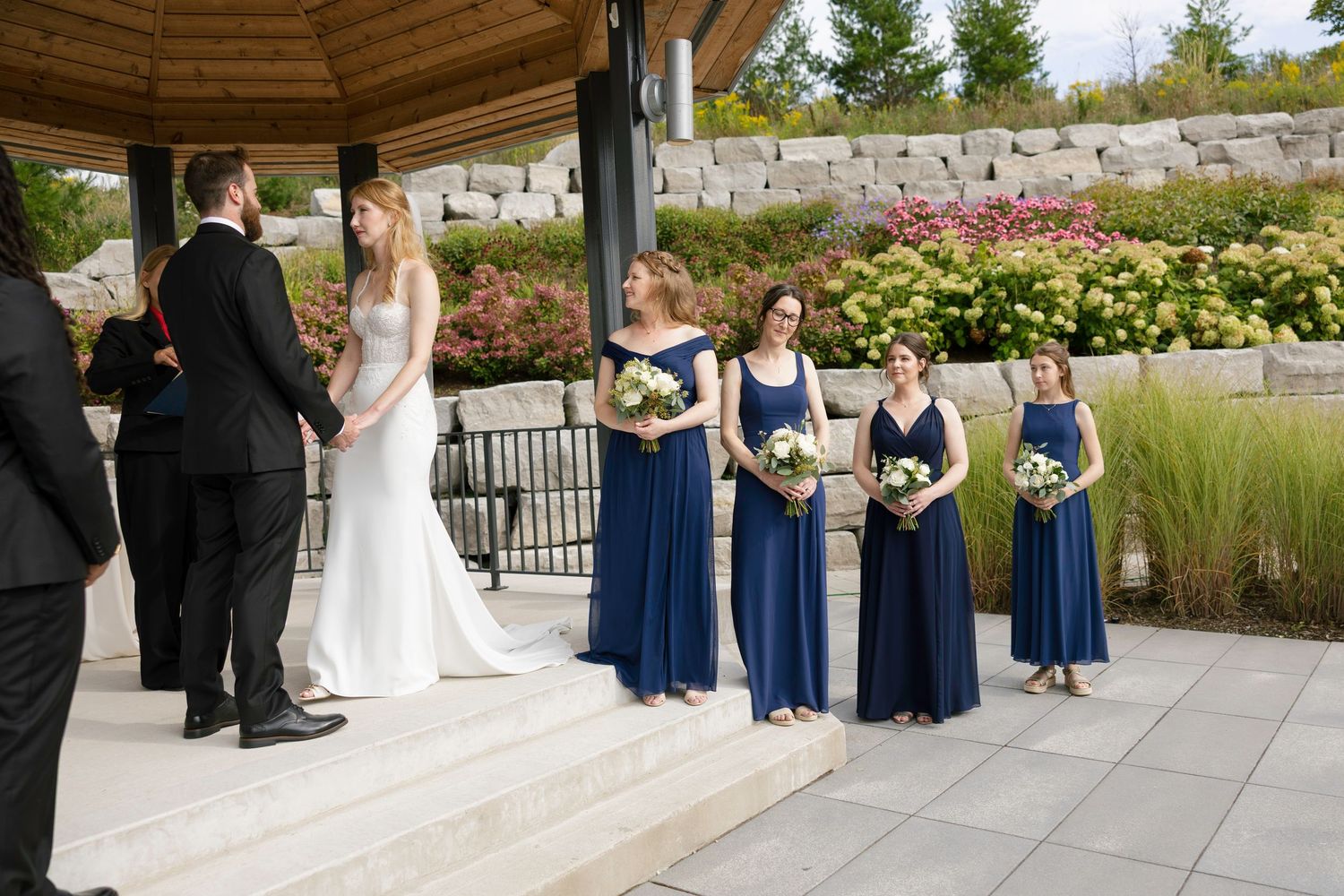 Wedding ceremony taking place at an outdoor gazebo with attendants in navy blue dresses holding bouquets.