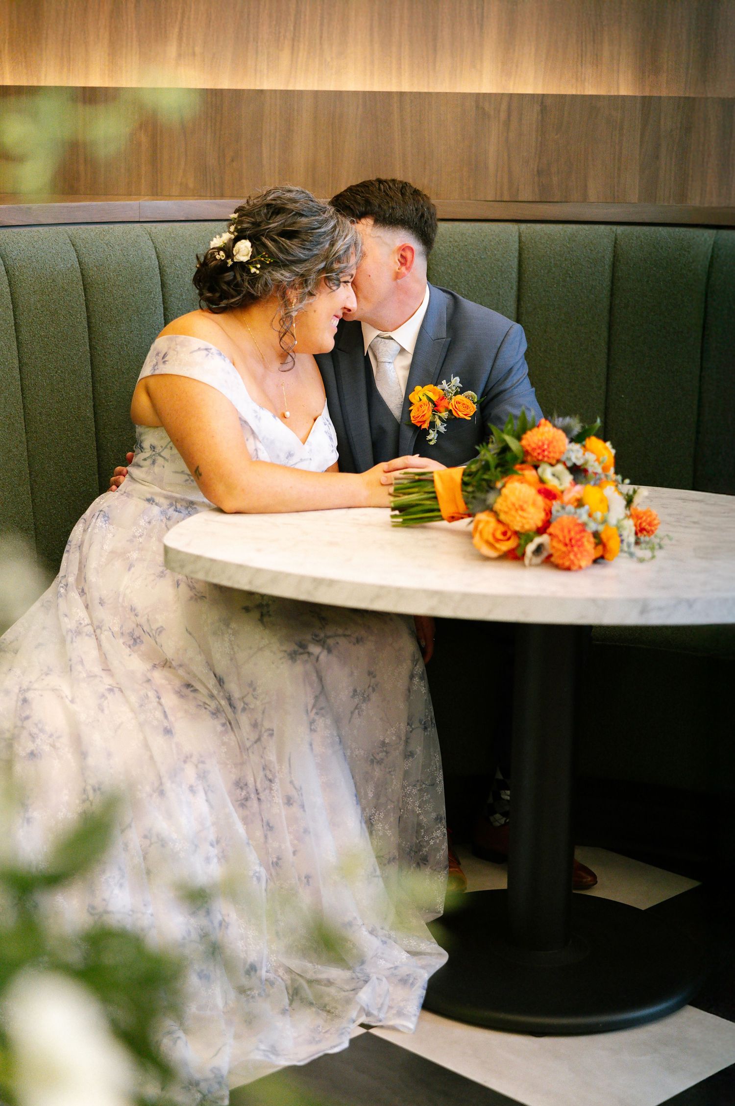 Bride and groom share an intimate moment in a green booth, with a vibrant orange floral bouquet on a marble table.