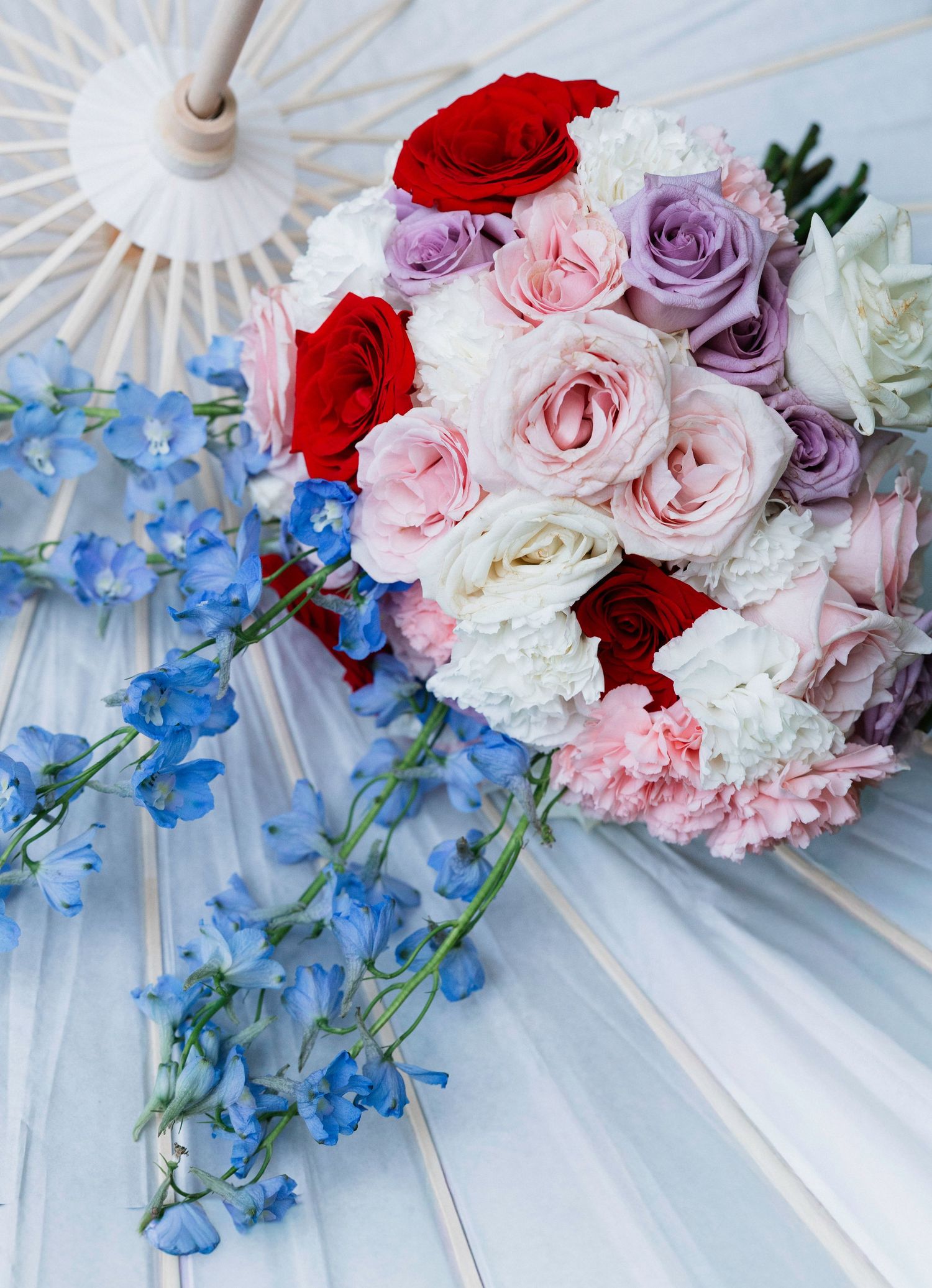 Elegant wedding bouquet with red, pink, lavender roses, white carnations, and blue delphinium on a white parasol.