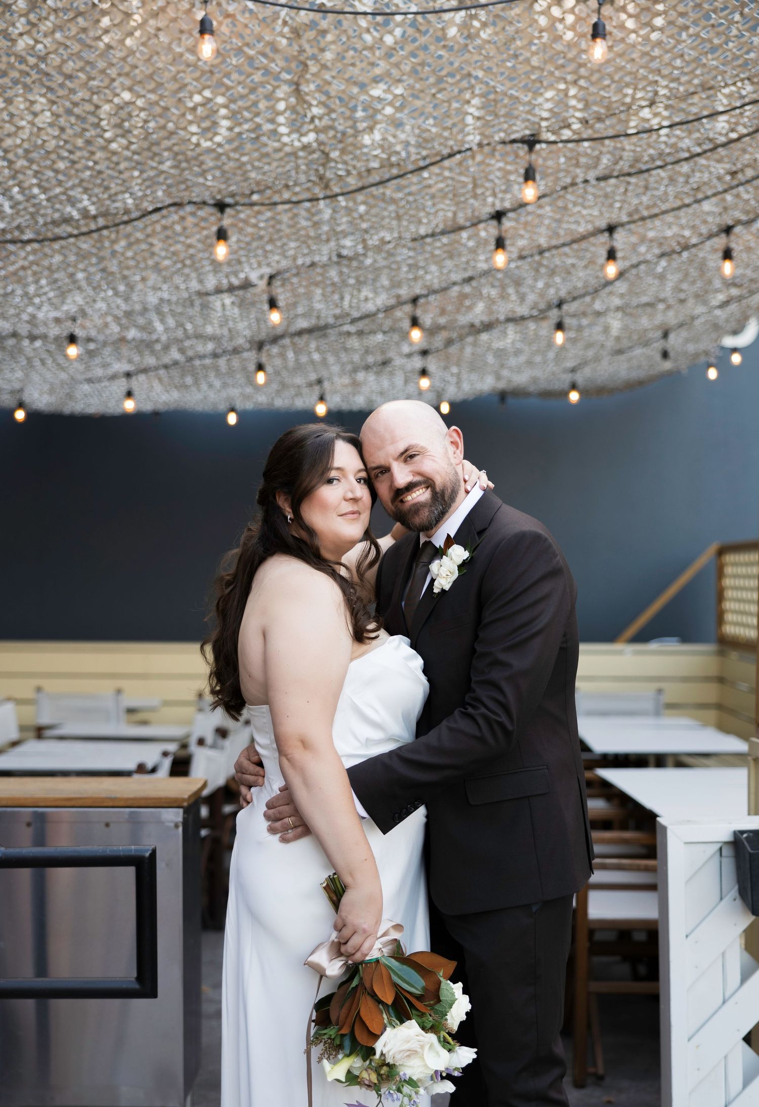 Bride and groom embrace under a sparkling chandelier with string lights in an elegant wedding venue.