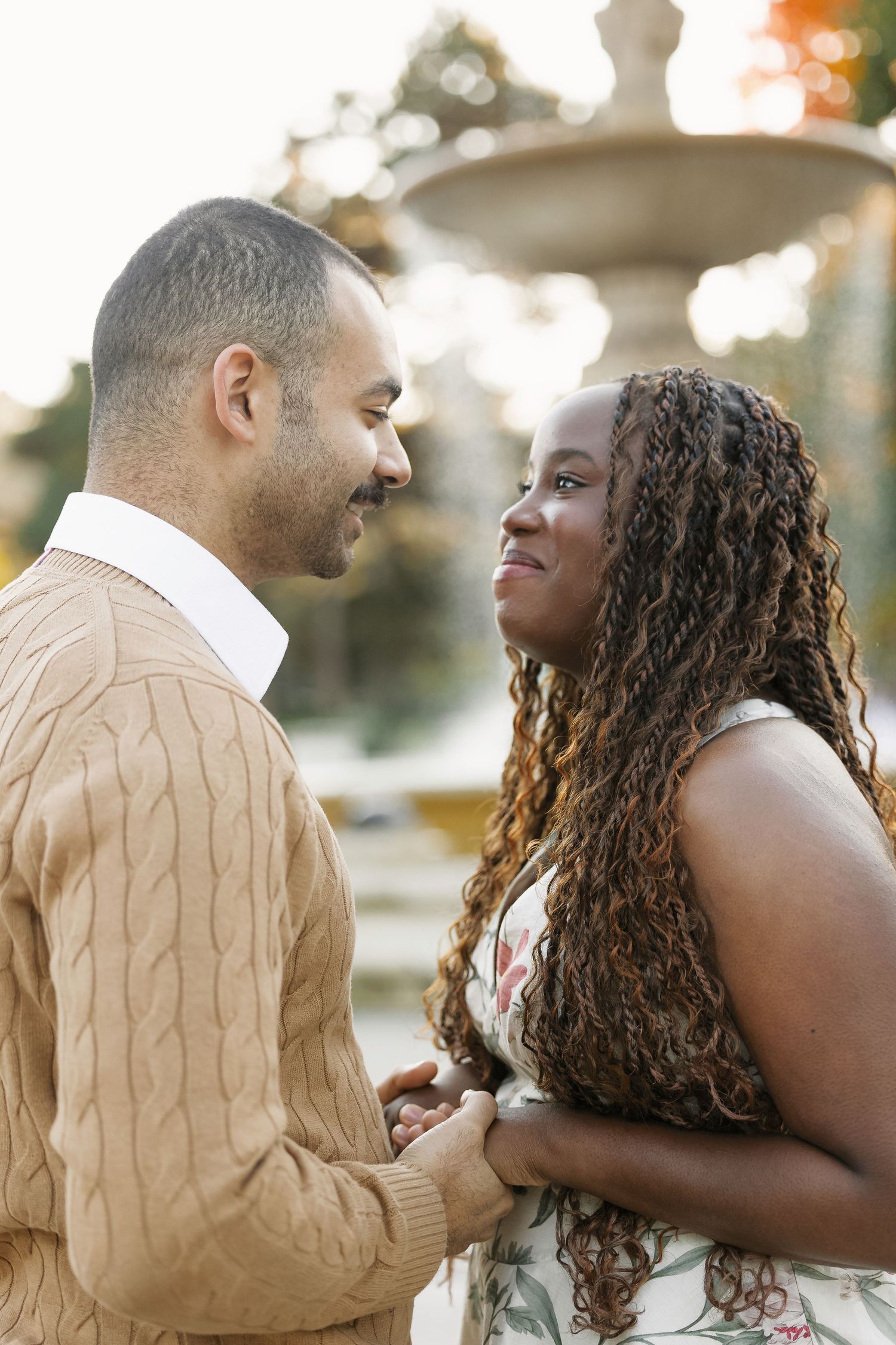 Interracial couple holding hands and smiling at each other near a fountain outdoors.