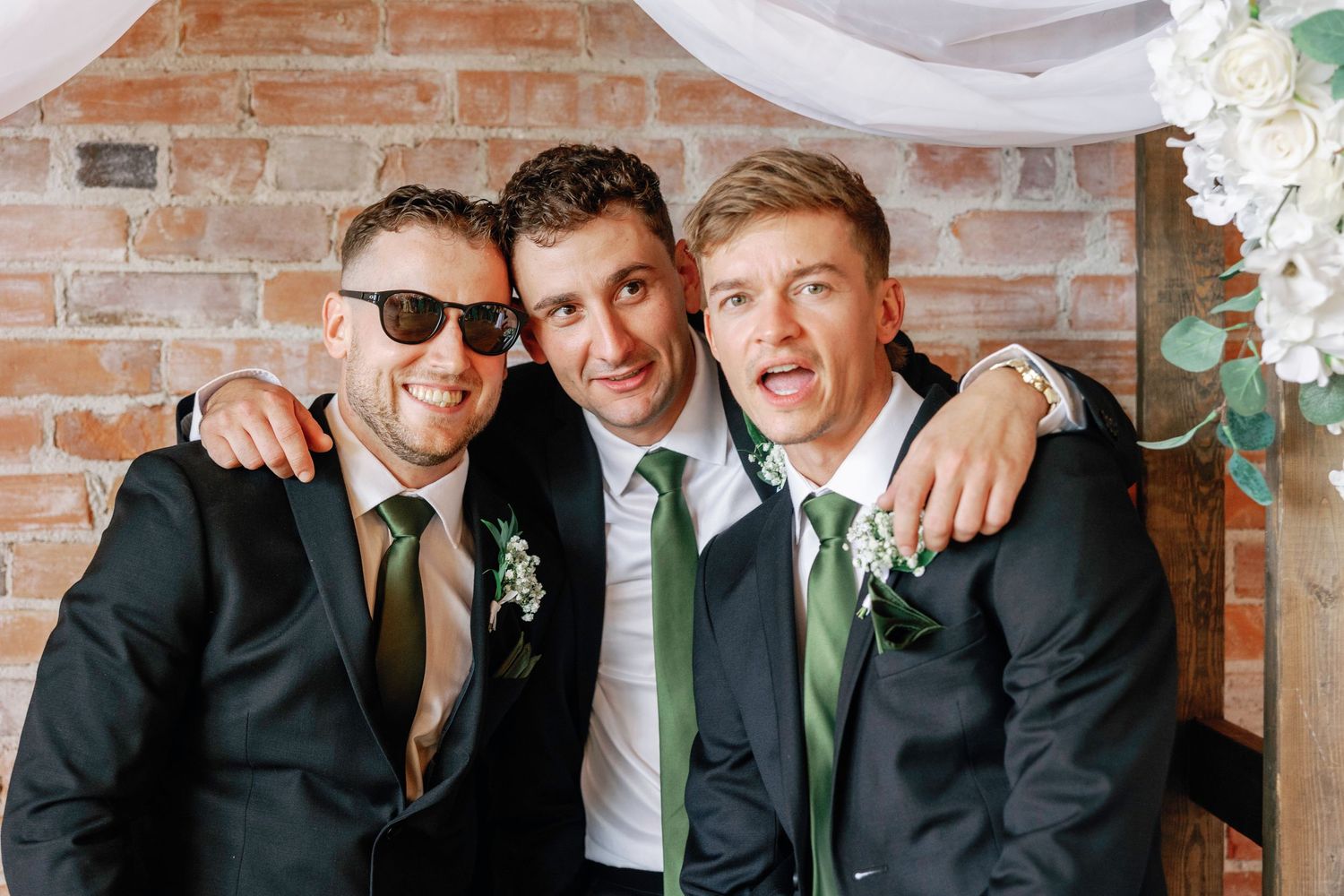 Groomsmen in black suits and green ties smile together against a rustic brick wall backdrop.