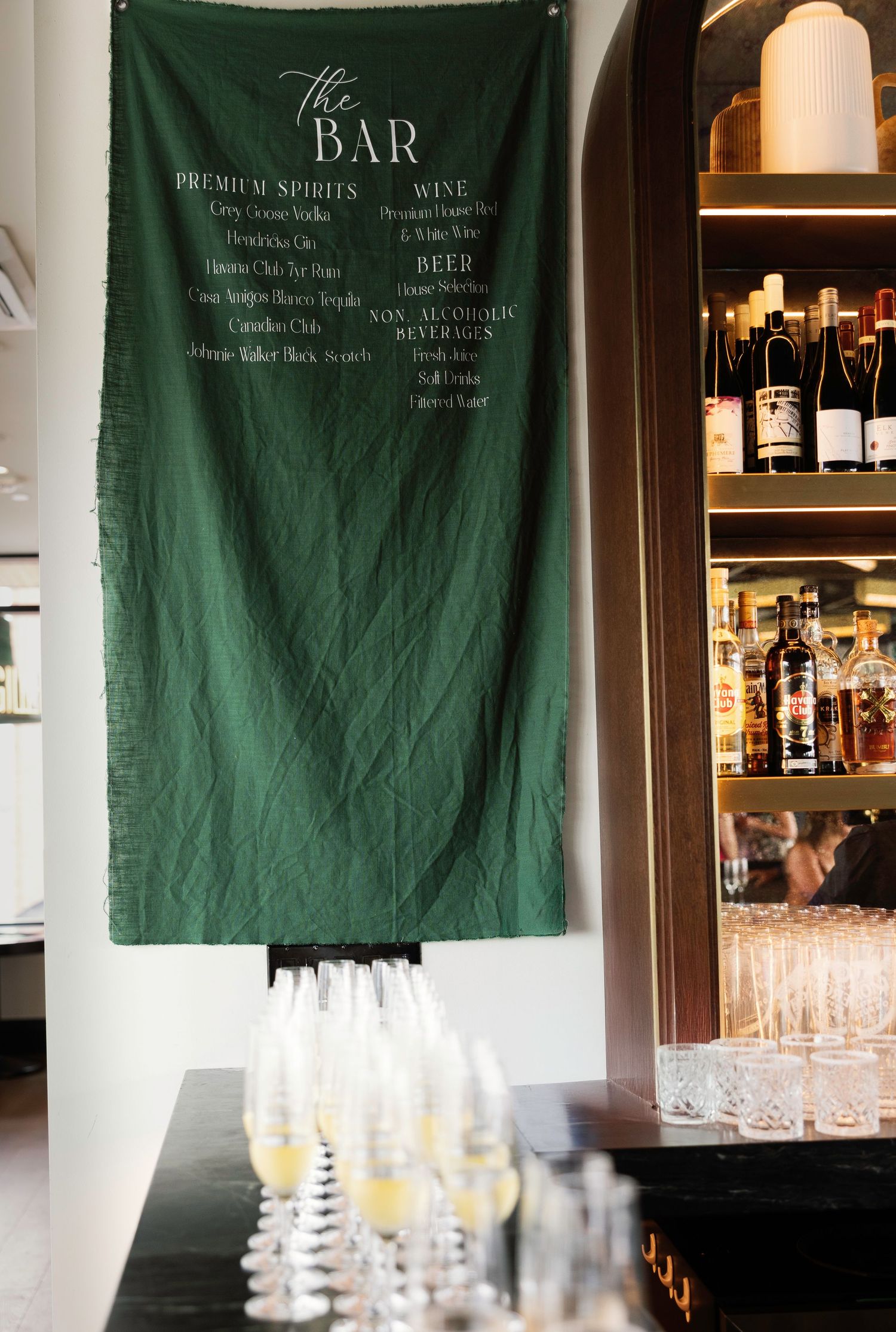Bar setup with green backdrop displaying drink menu and glassware for wedding reception.