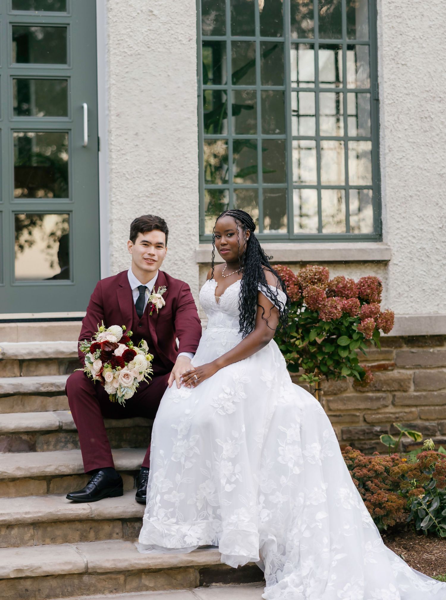 A couple in wedding attire shares romantic moments on a porch with white columns and burgundy floral decorations.