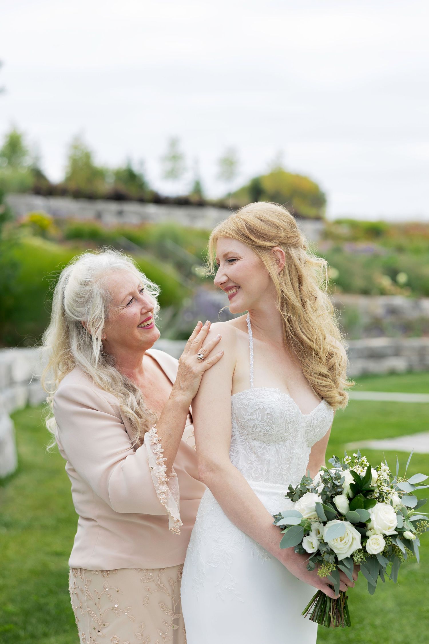 A bride and companion share a joyful moment outdoors, laughing together while holding a white floral bouquet.