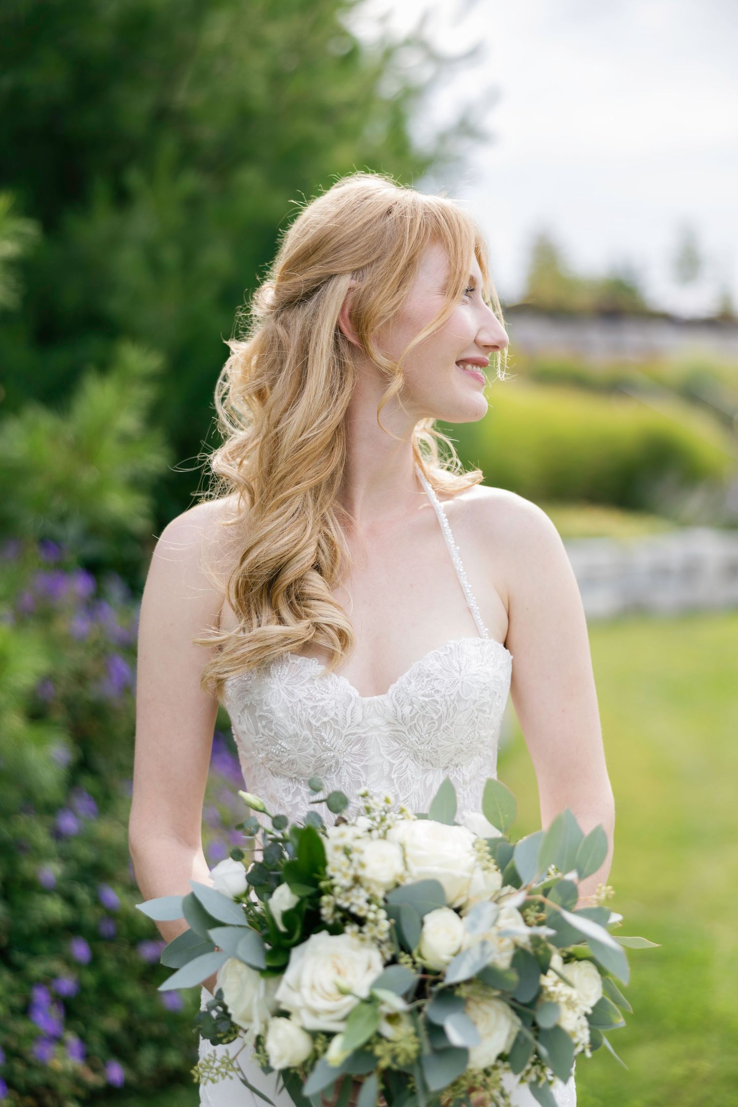 A bride in a white wedding dress holds a lush bouquet of white flowers and eucalyptus against a green garden backdrop.