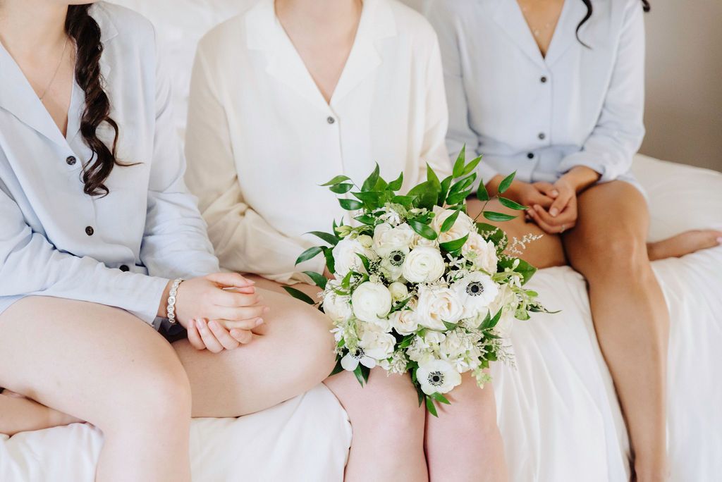 Bridesmaids in soft pink dresses sit together holding white floral bouquets during a wedding celebration.