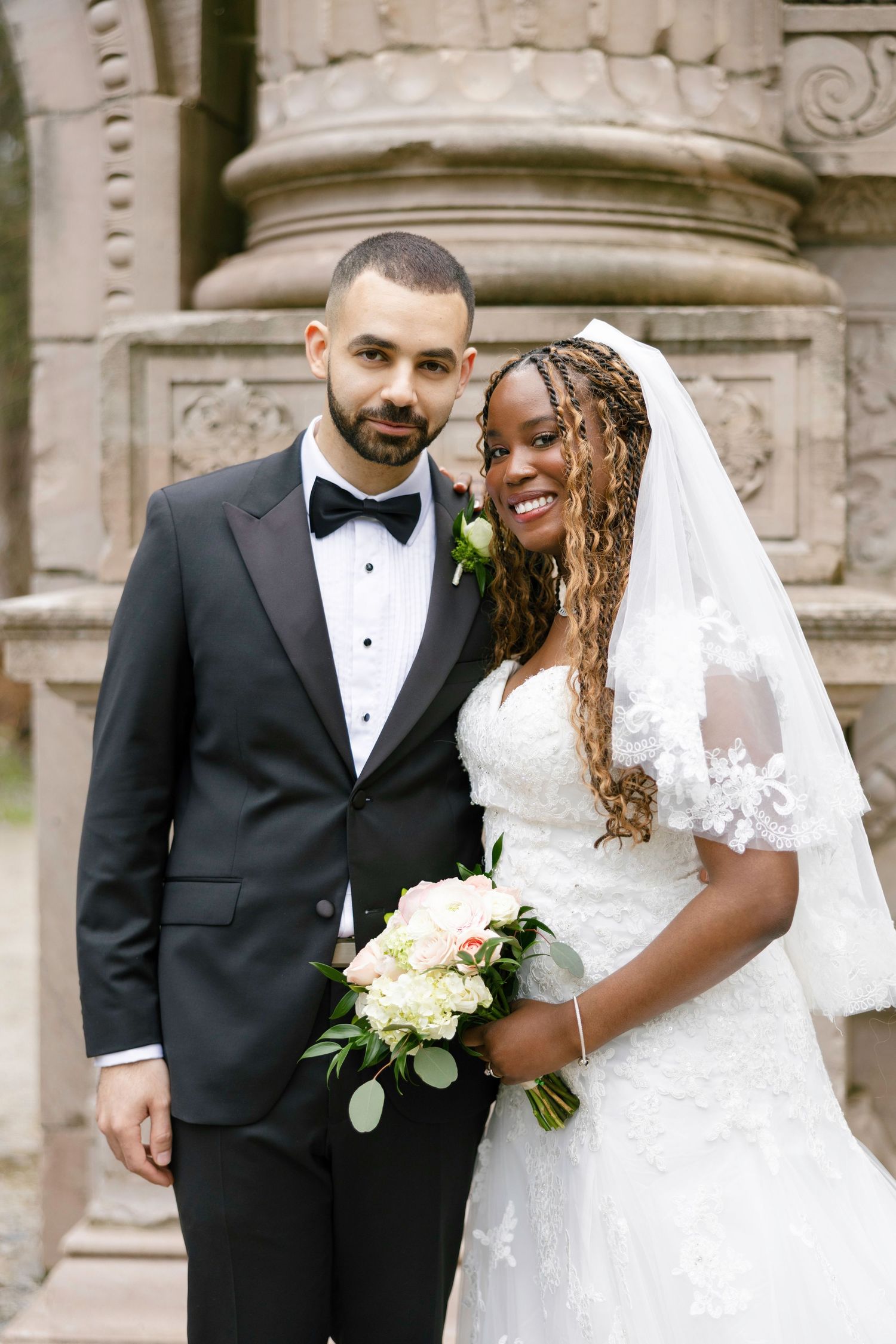 Interracial couple posing in wedding attire, groom in black tuxedo, bride in lace gown holding white bouquet.