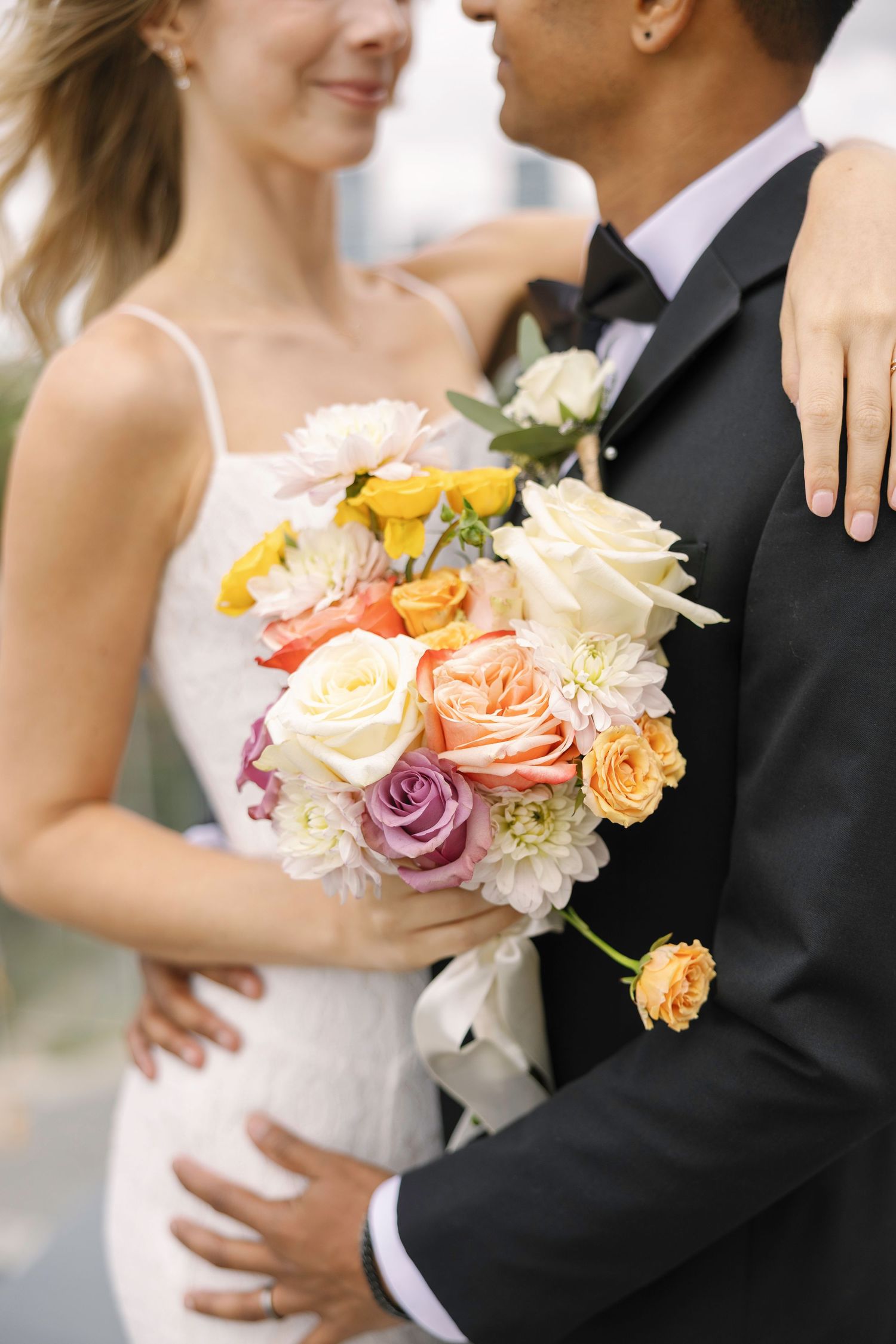 Bride and groom embrace holding a colorful wedding bouquet with roses and dahlias in peach, yellow, and purple hues.