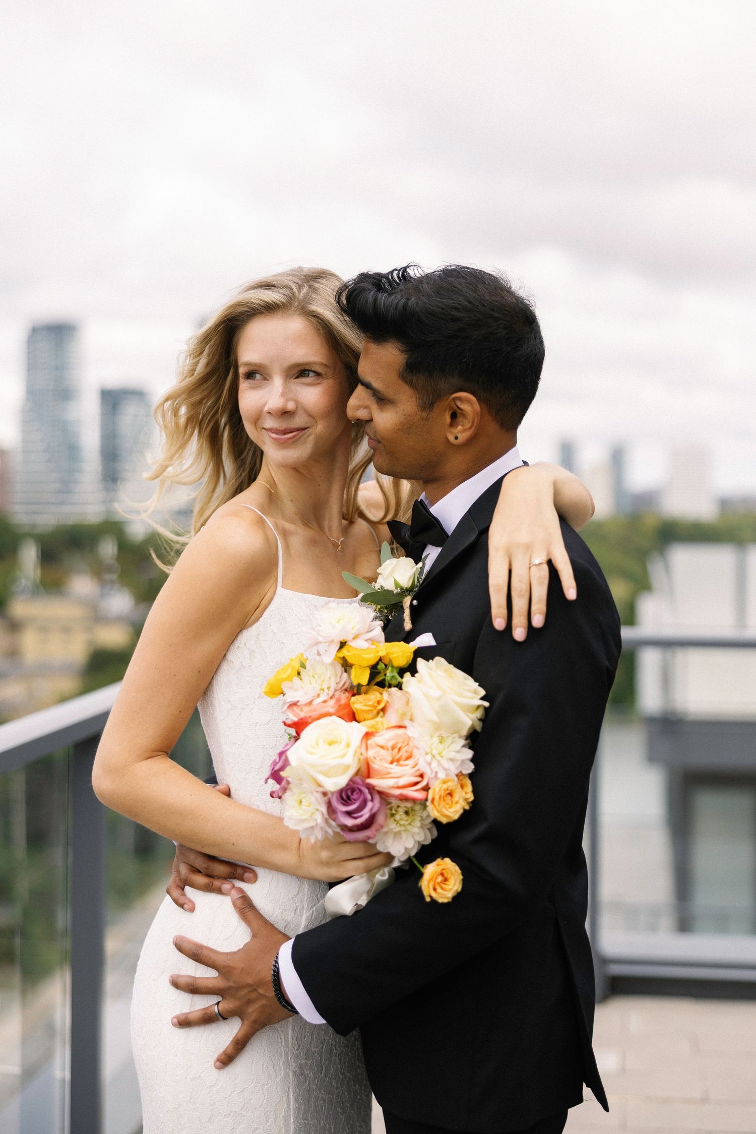Bride and groom embrace on rooftop with city skyline, bride holds colorful bouquet in white gown, groom in black tuxedo.