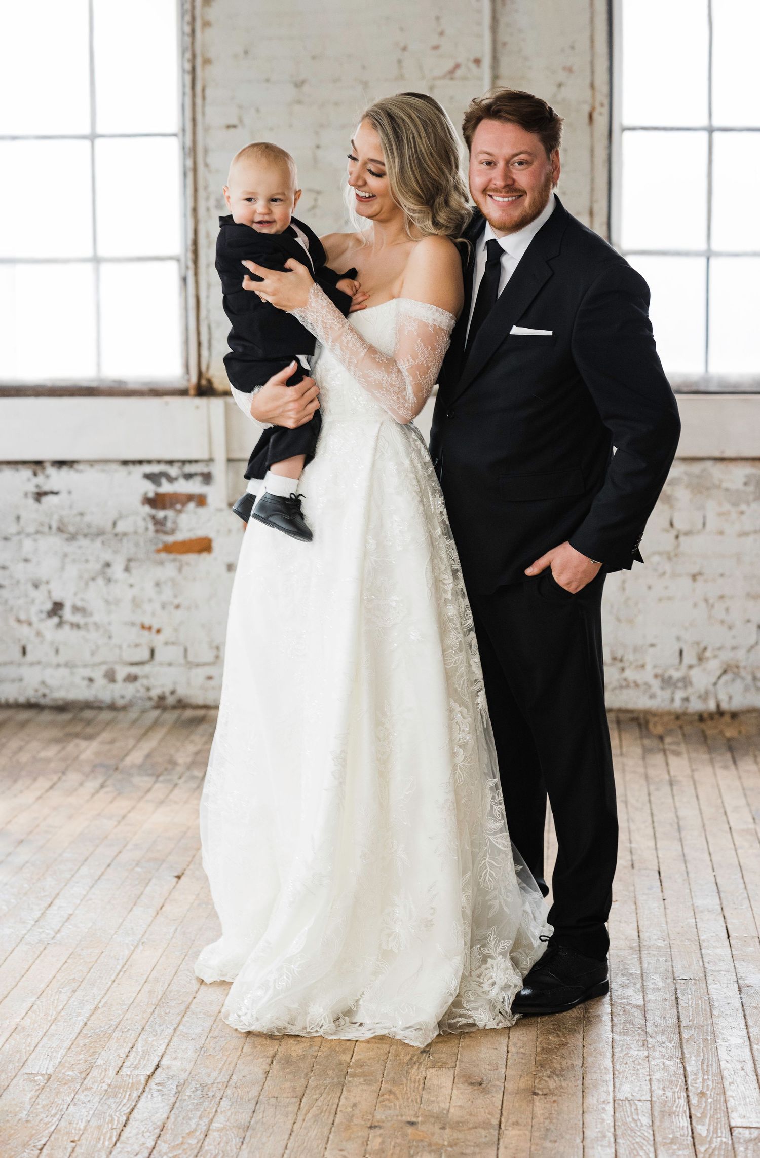 A bride in white dress and groom in black suit share a tender moment with a baby at an indoor wedding venue.