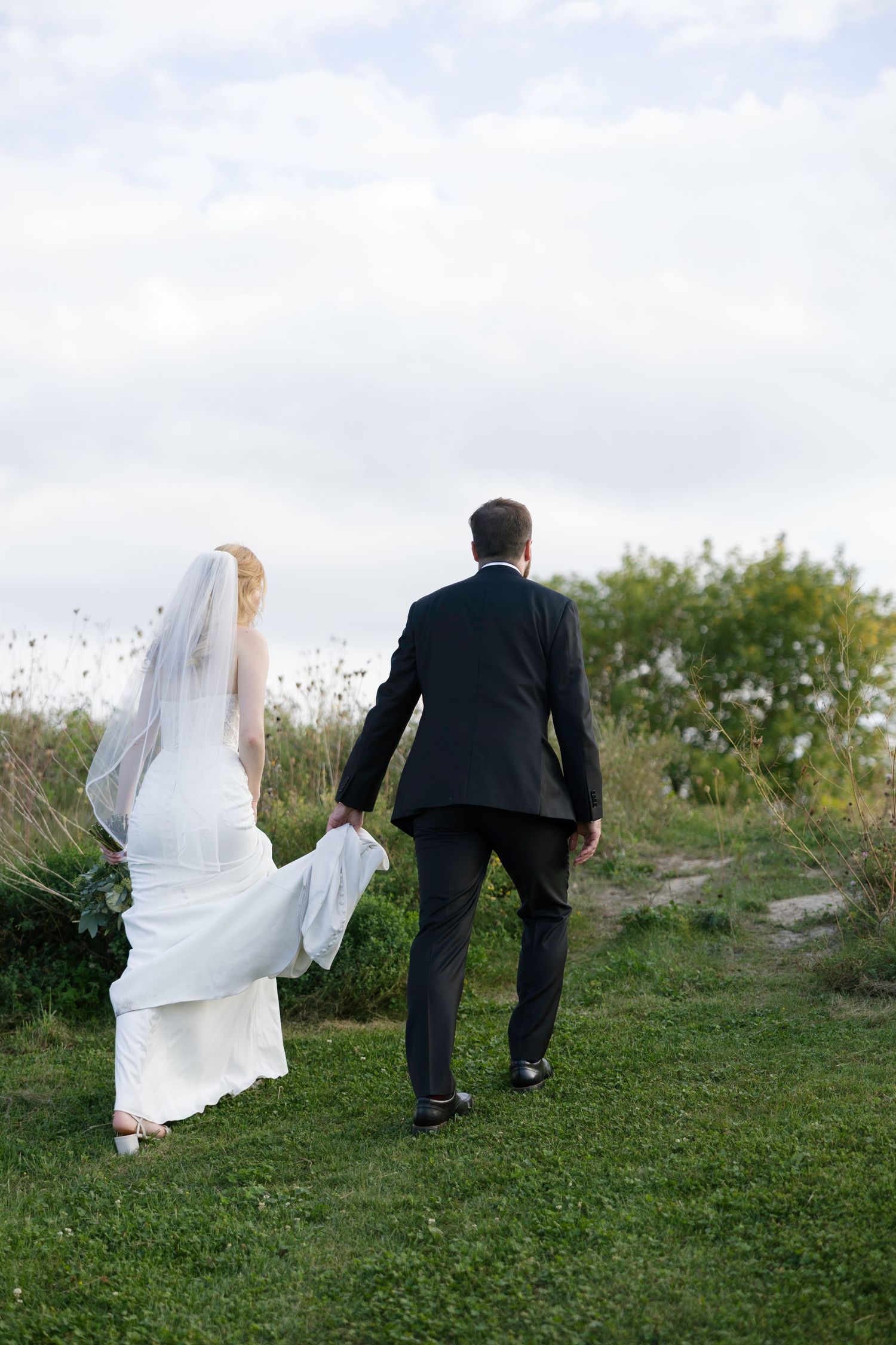 A couple in wedding attire walks up a grassy hillside as the bride's white veil flows in the wind.
