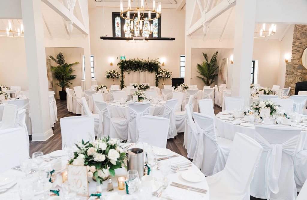 Elegant white wedding reception venue with floral centerpieces, draped chairs, and crystal chandeliers in a vaulted ceiling room.