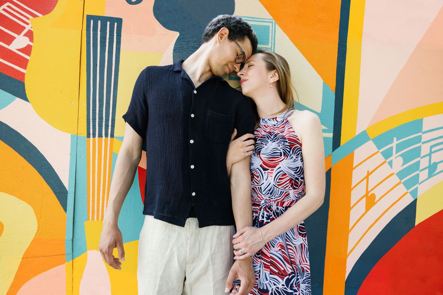 Couple poses playfully against colorful geometric mural wall in urban setting.
