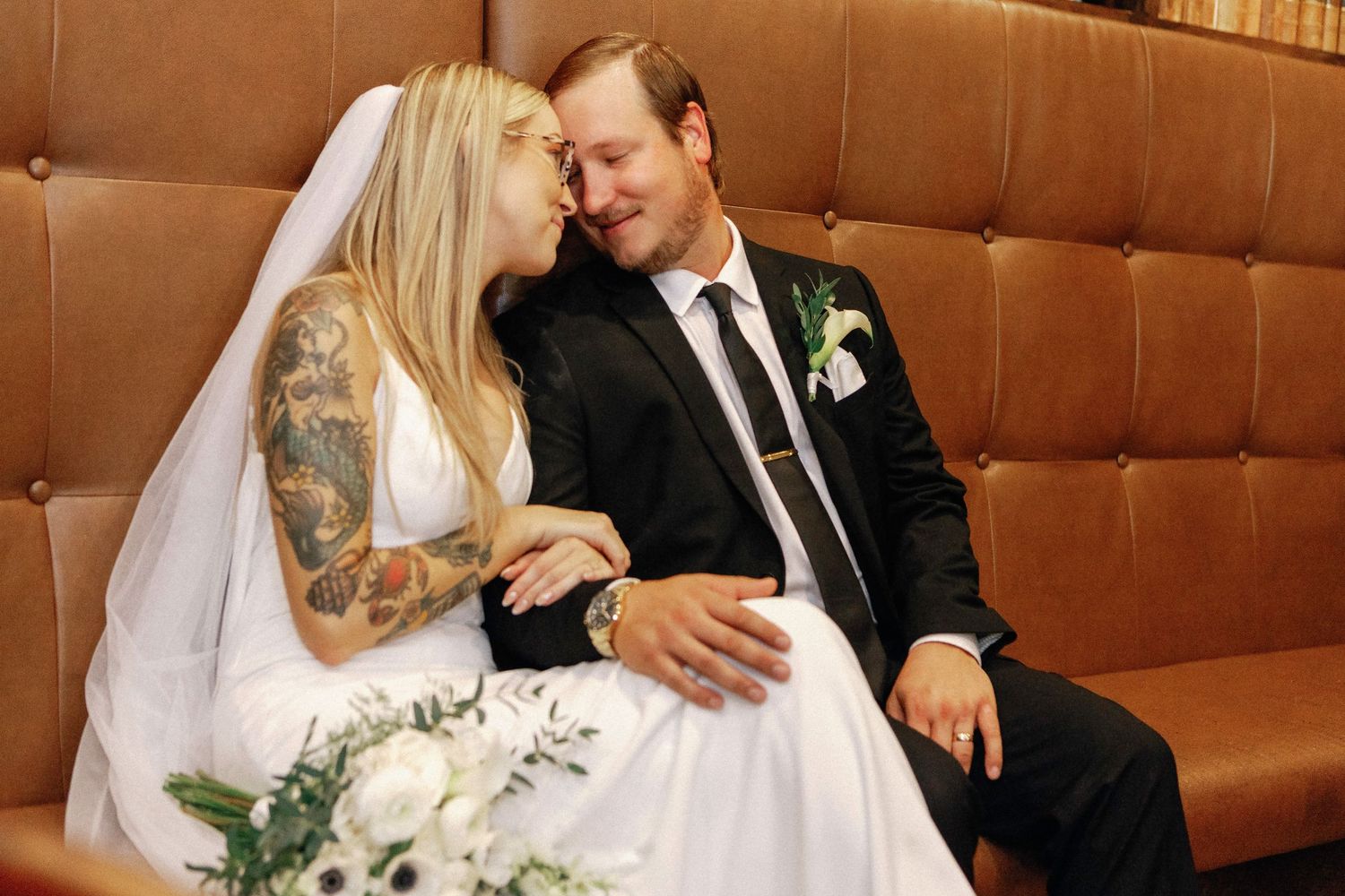A bride and groom share an intimate moment while sitting together in a brown leather booth at their wedding reception.