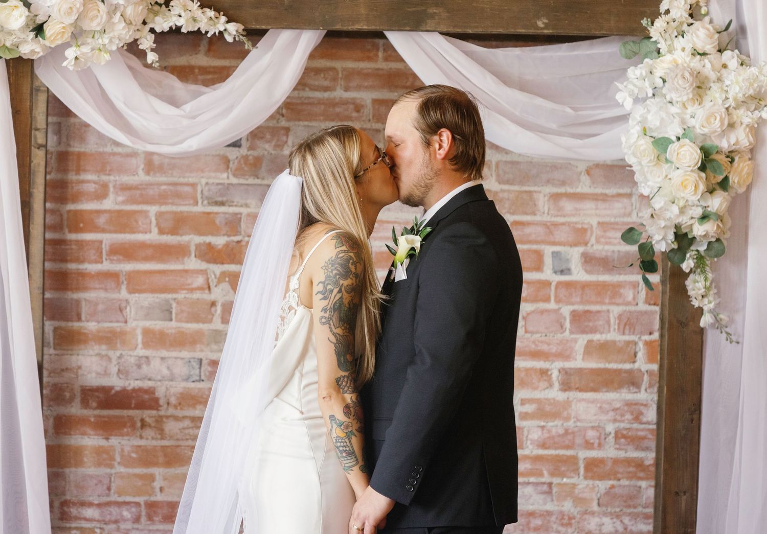 Wedding altar decorated with white draping and floral arrangements against brick wall backdrop.