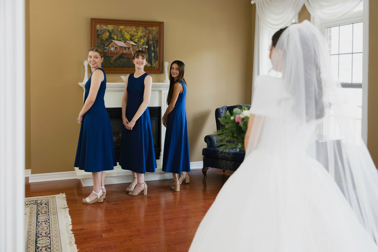 Bride in white gown and veil stands before three bridesmaids in navy blue dresses in an elegant home.