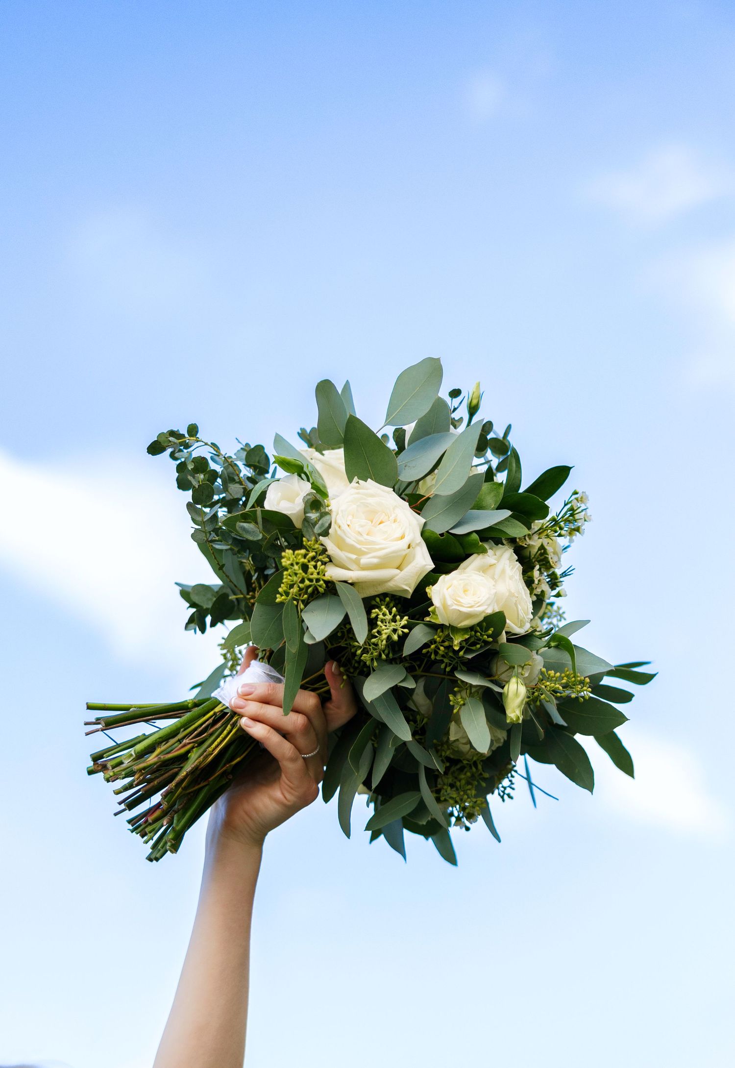 A hand holds a bridal bouquet of white roses and eucalyptus greenery raised against a bright blue sky.