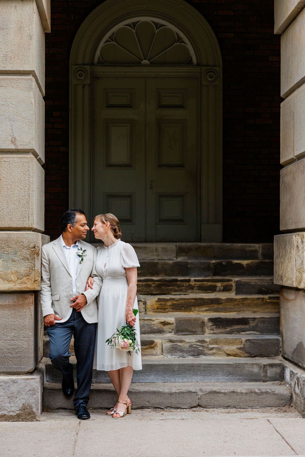 Couple in wedding attire poses on stone steps before elegant arched door, bride holding pink bouquet.