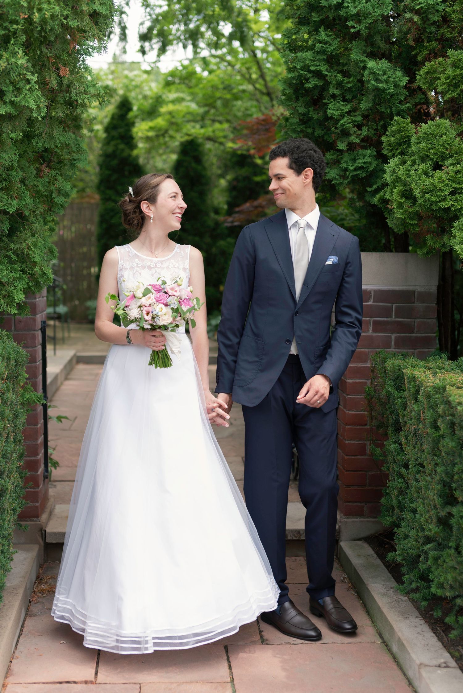 Wedding couple poses on garden pathway with brick walls and green foliage.