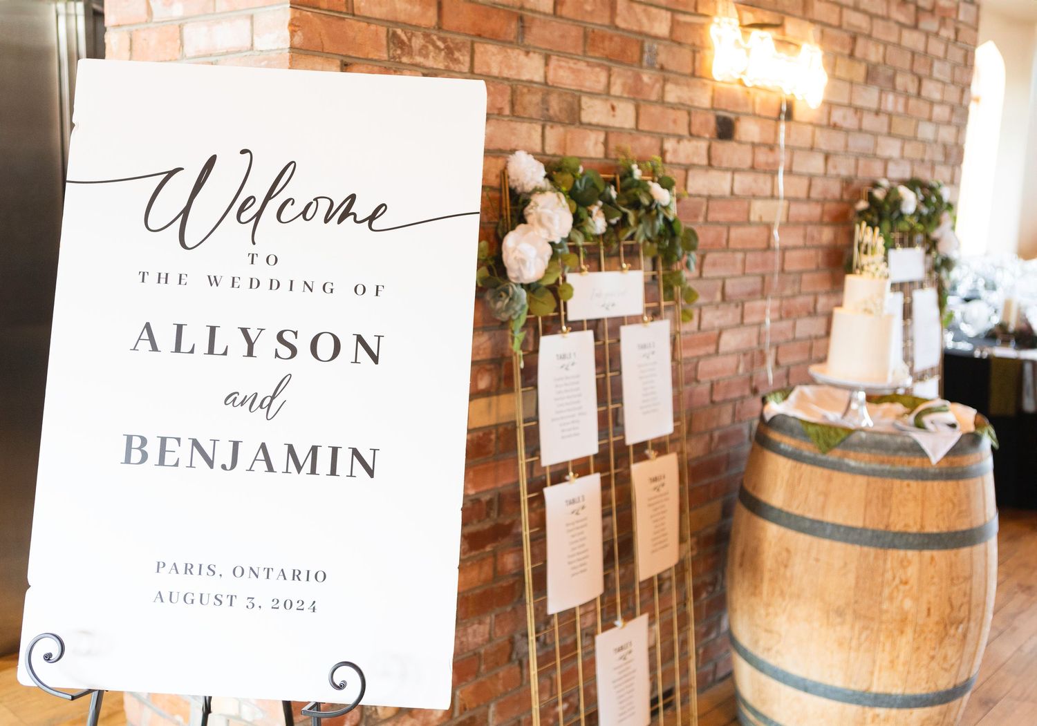 Wedding welcome sign and rustic wine barrel decor against exposed brick wall with floral arrangements.