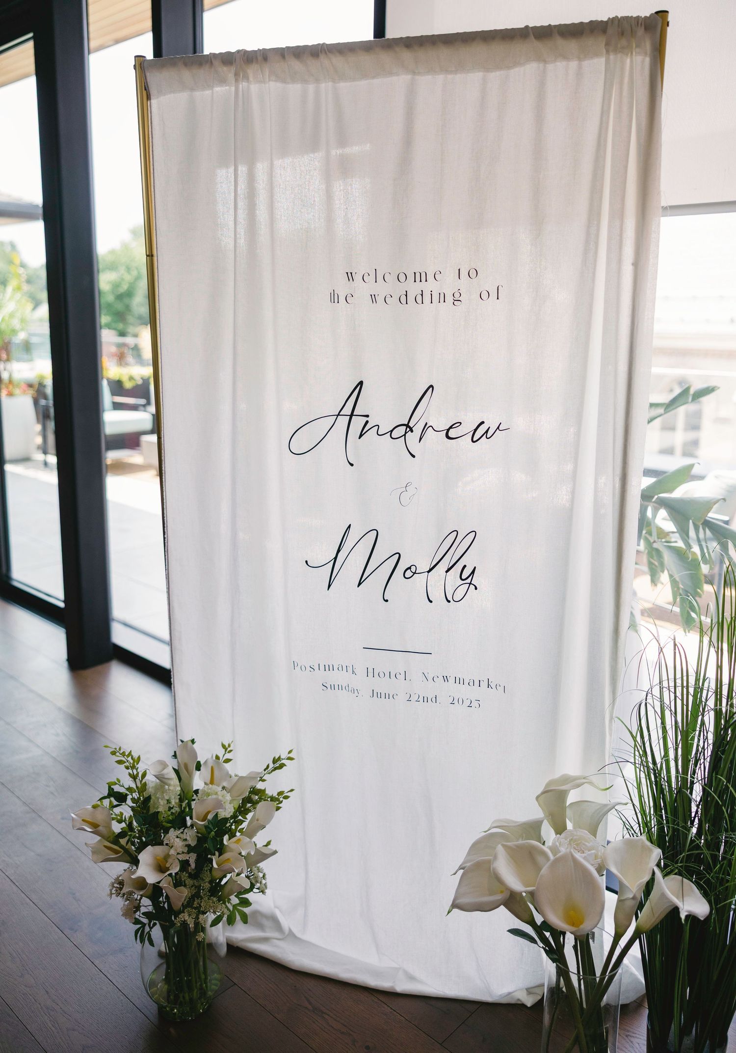 Elegant wedding welcome sign with script lettering displayed on white fabric backdrop with floral arrangements.
