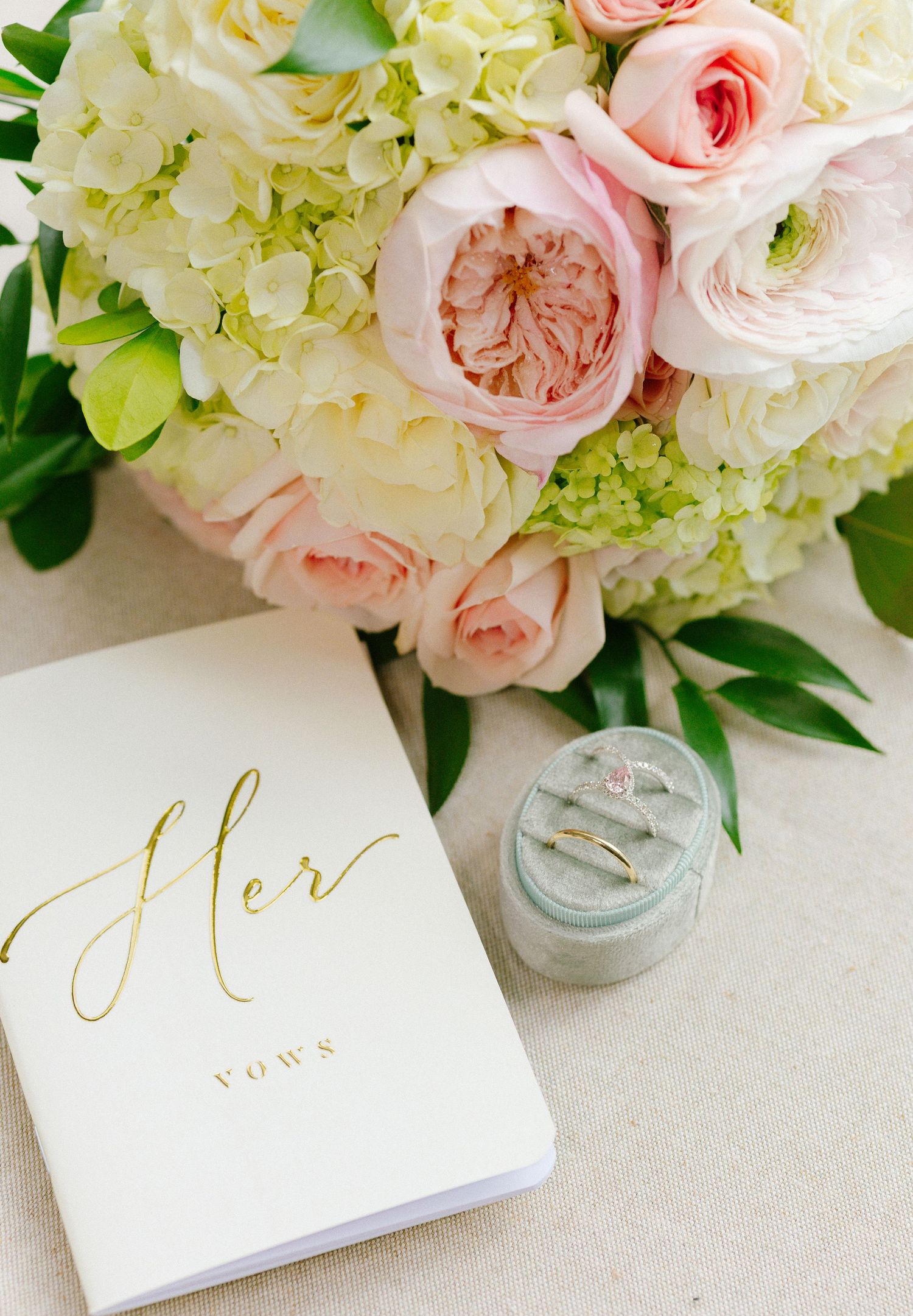 Elegant wedding flat lay with pink roses, hydrangeas, a gold Her Vows book, and a diamond ring in a velvet box.