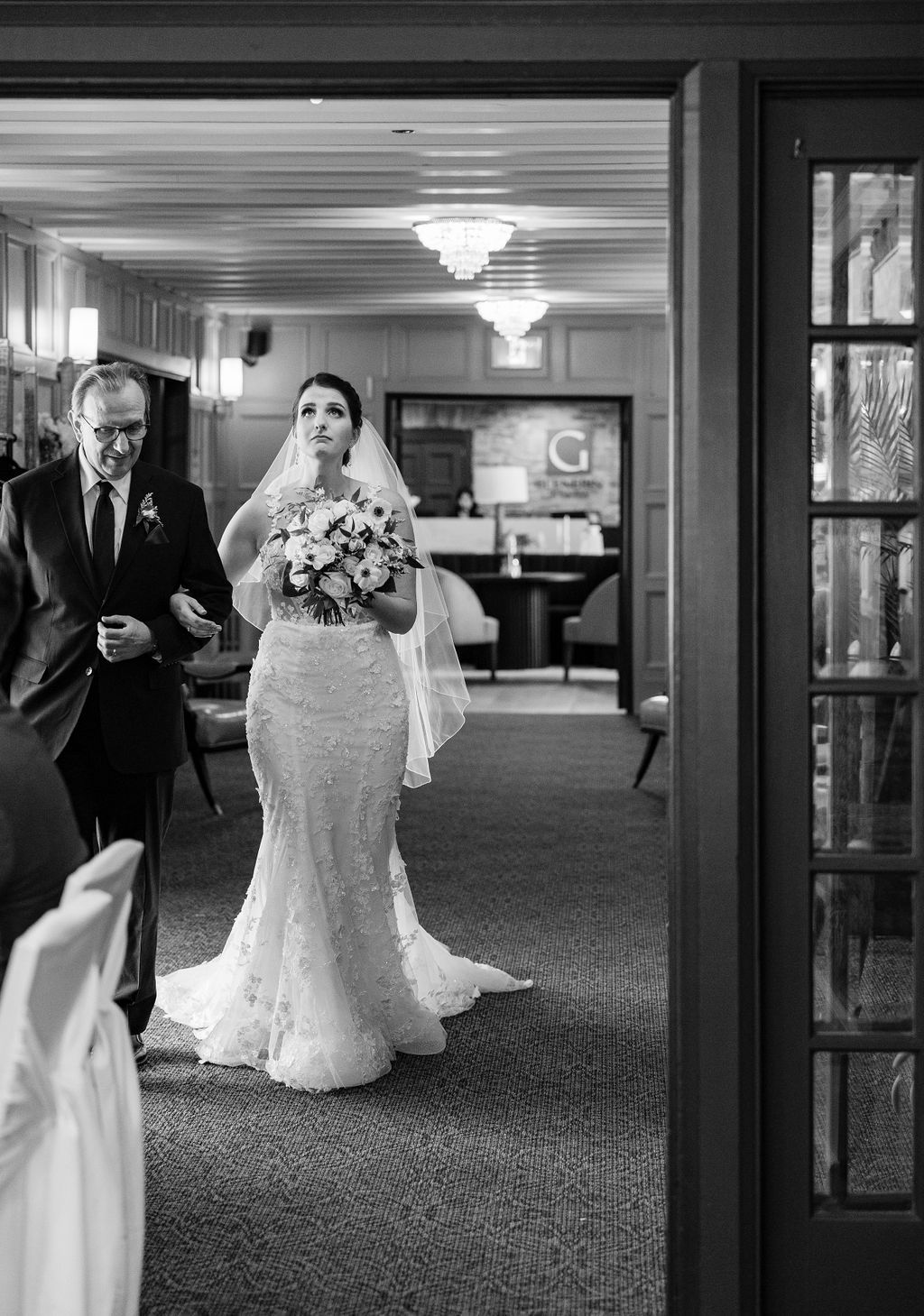 A black and white wedding photograph showing a bride and groom walking down a carpeted hallway.