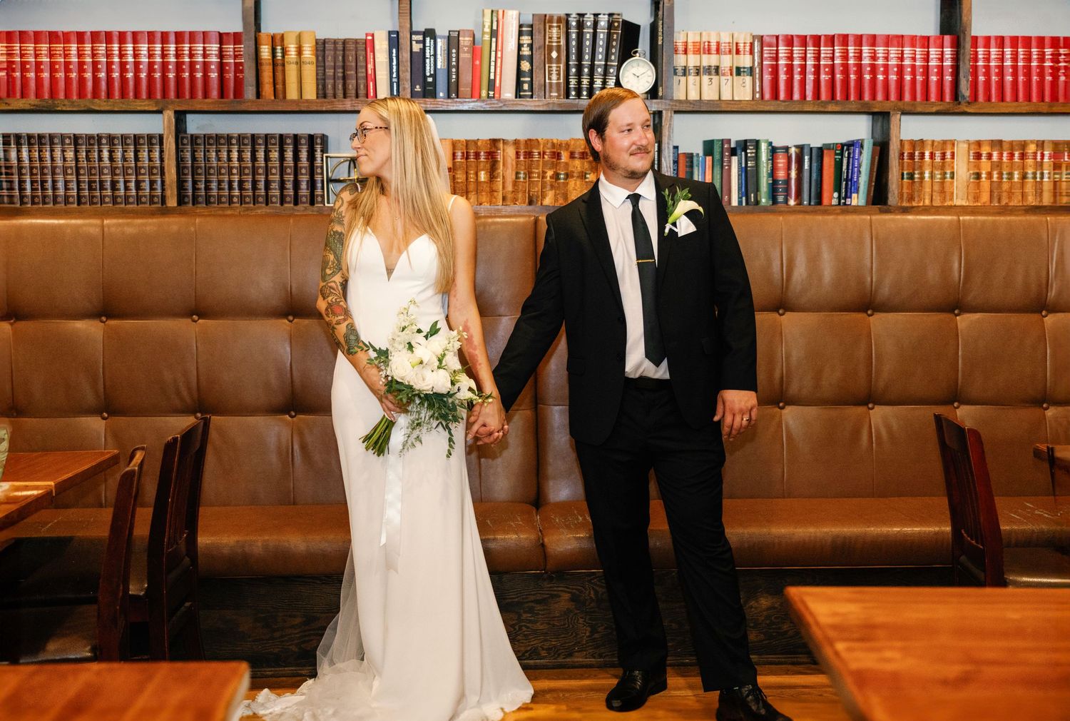 A couple in wedding attire walks through a rustic venue with wood paneling and leather booth seating.
