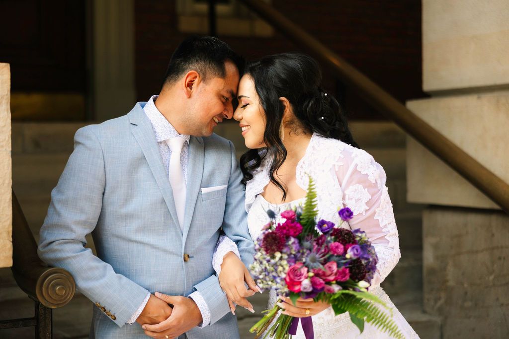 A bride and groom share a romantic forehead touch, she holds a purple bouquet, he wears a light gray suit.