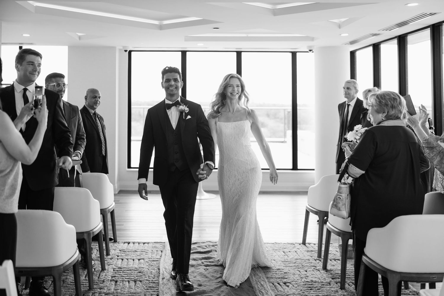 Bride and groom walk hand in hand down the aisle in a modern venue, black and white wedding photo.