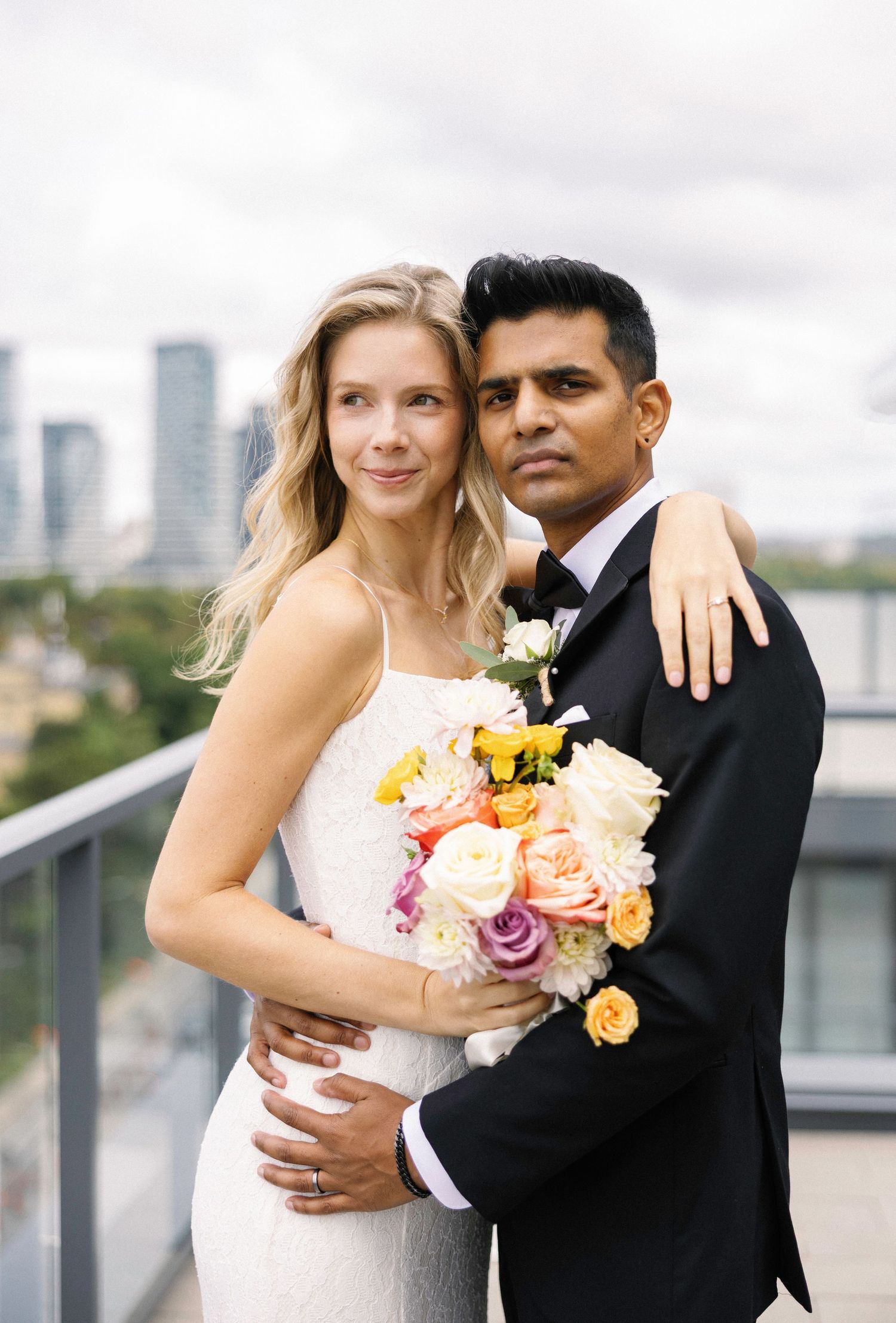 Bride and groom pose on rooftop terrace, she holds colorful bouquet, city skyline backdrop, wedding portrait.