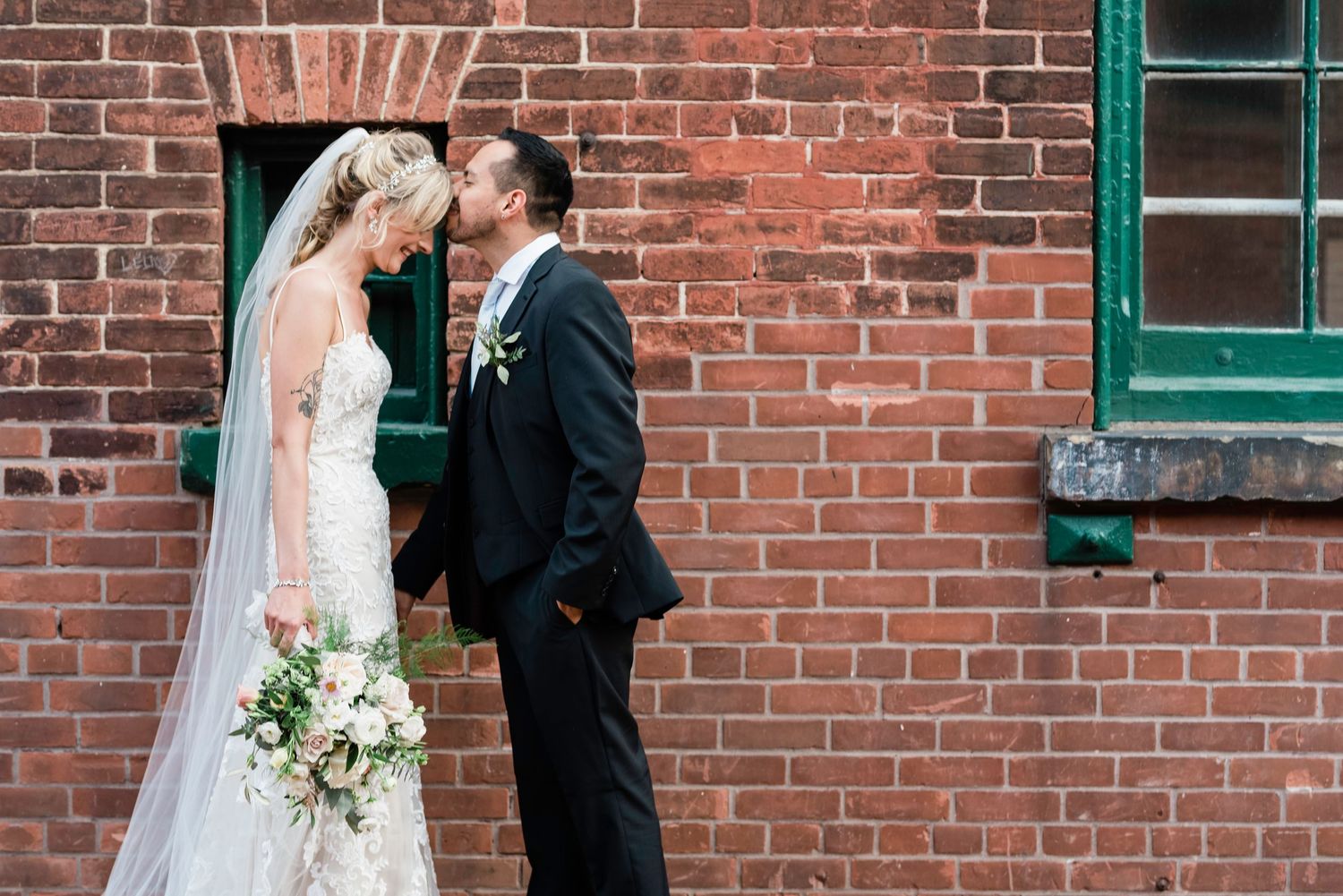 Bride and groom sharing a kiss against a rustic red brick wall, bride holding a white floral bouquet with veil.