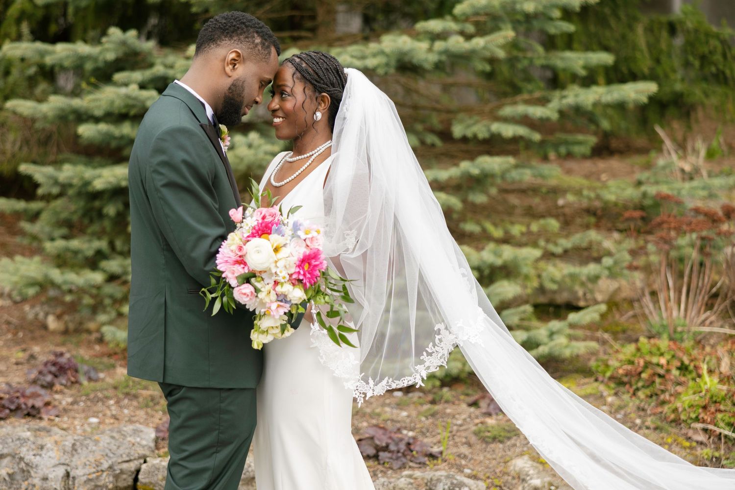 Bride and groom in elegant wedding attire share a romantic moment outdoors, bride holding a vibrant pink and white bouquet.