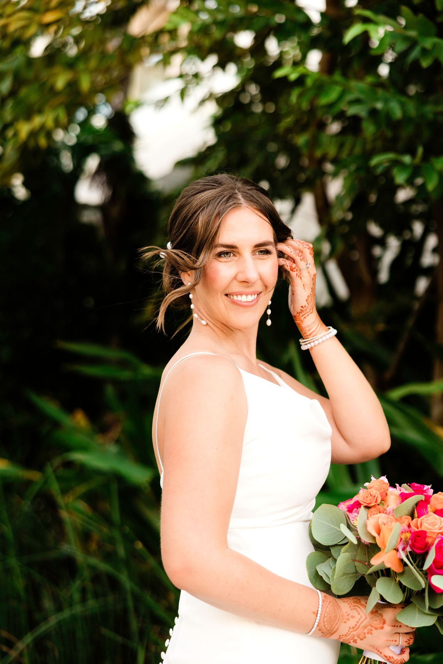 A person in a white wedding dress holds a colorful bouquet while posing outdoors among lush greenery.