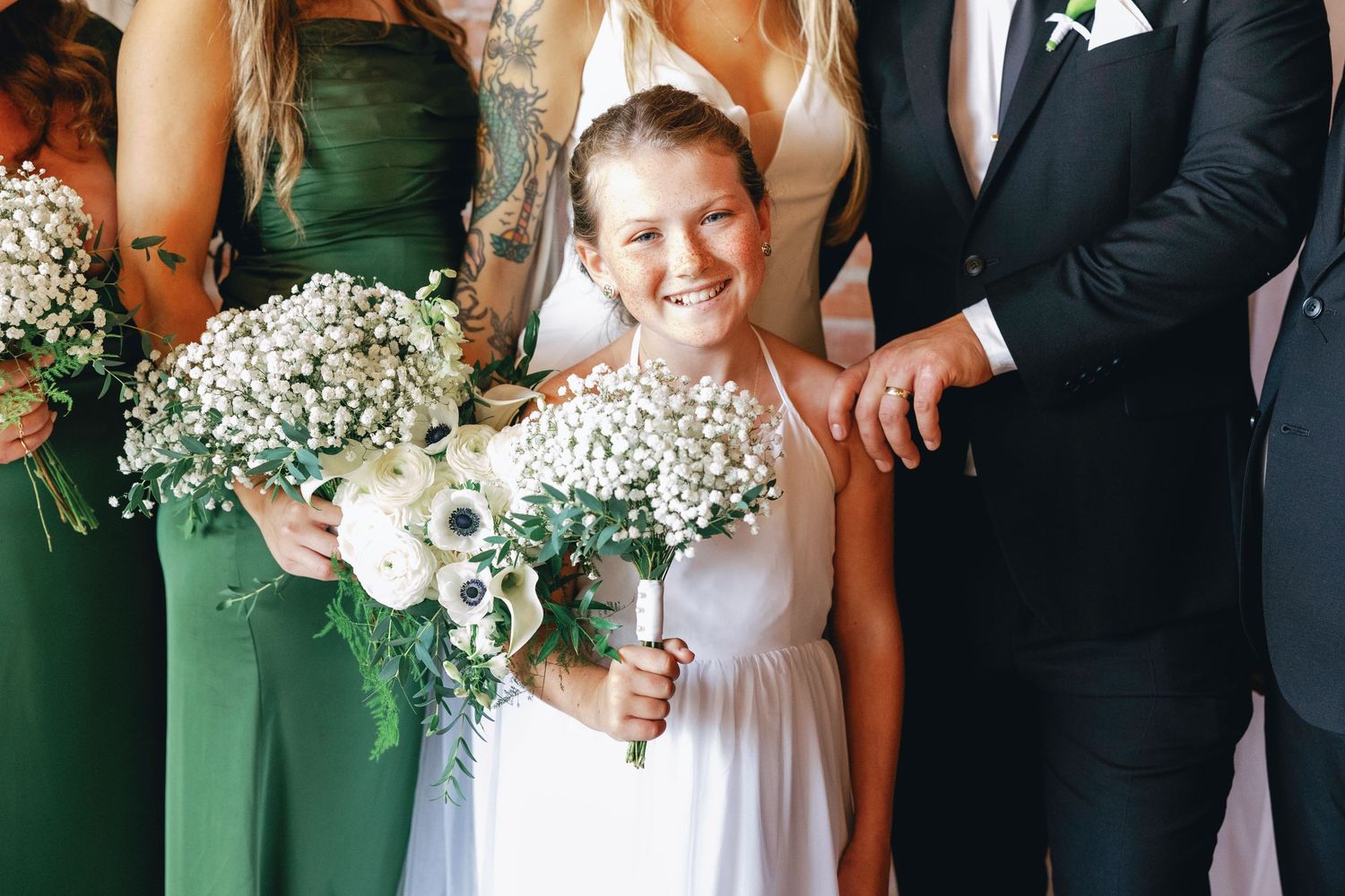 Wedding party members in green dresses and black suits pose together holding white floral bouquets.