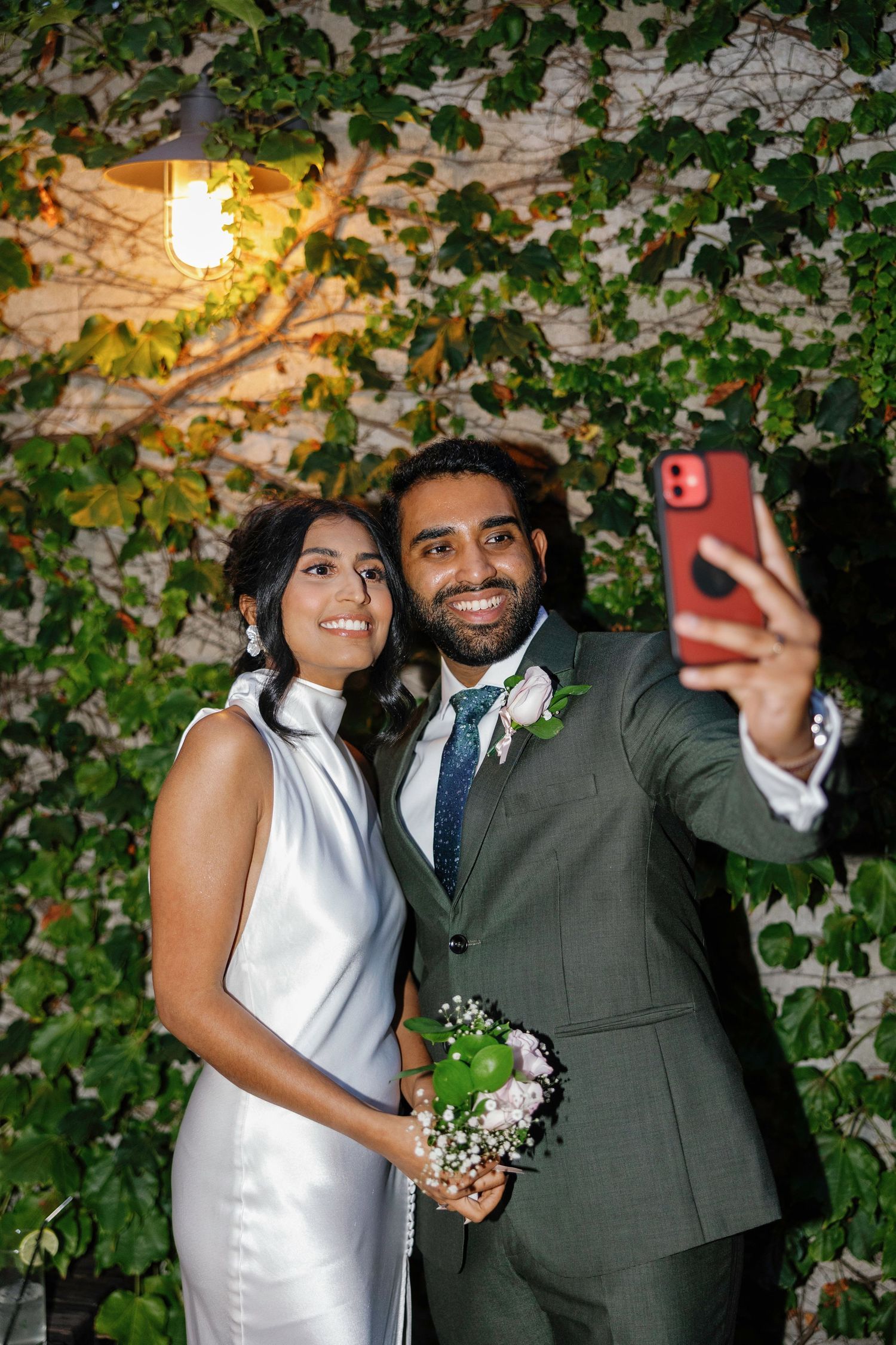 Newlyweds take romantic selfie against ivy-covered wall with evening lighting.