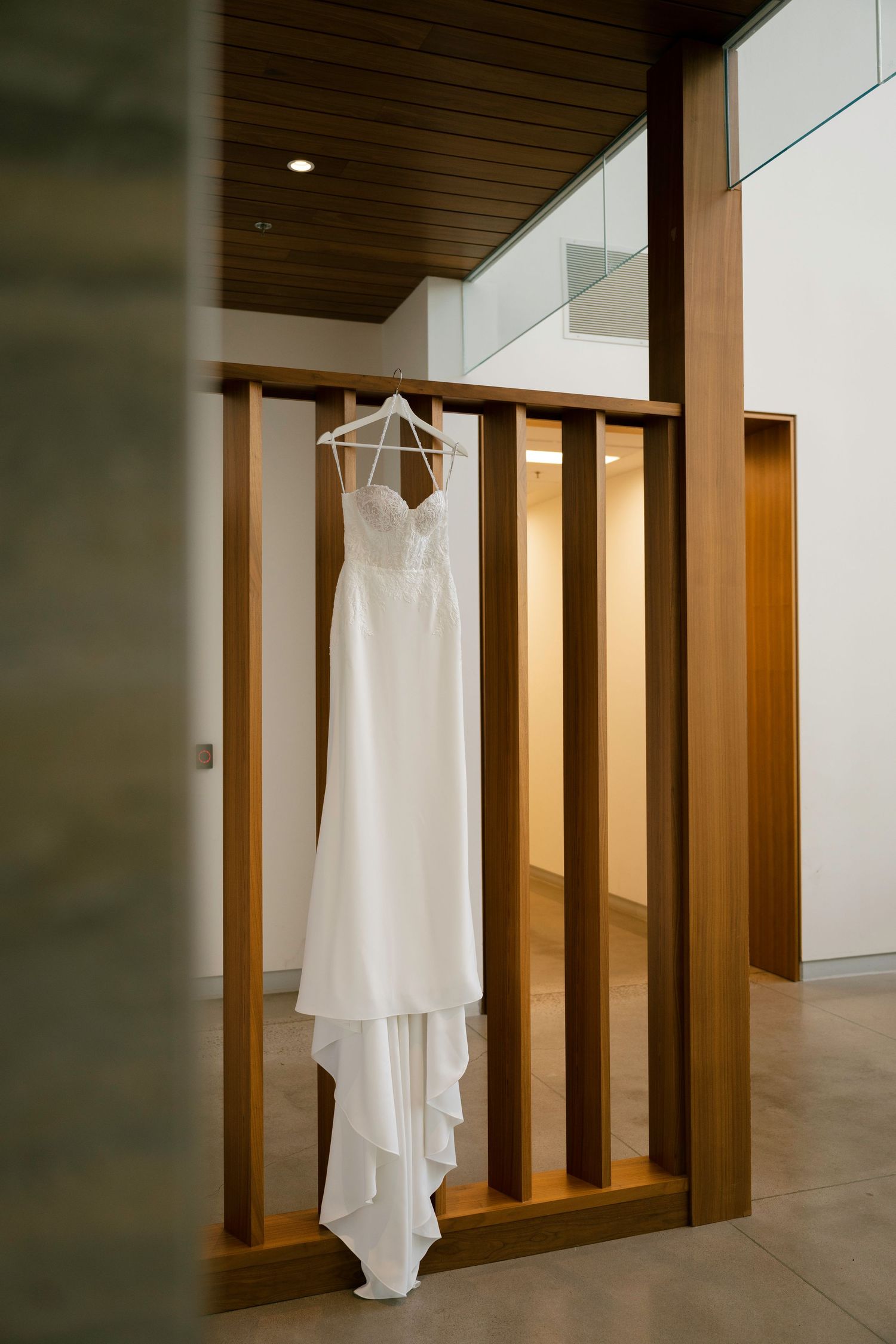 A white wedding dress hangs elegantly against a modern wooden wall with warm lighting in a minimalist hallway setting.