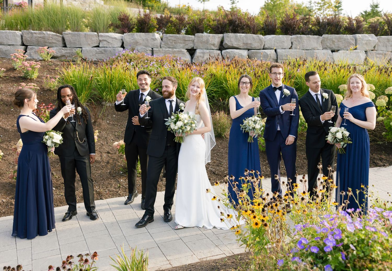 Wedding party in formal attire poses outdoors against stone wall with blooming wildflowers and greenery.