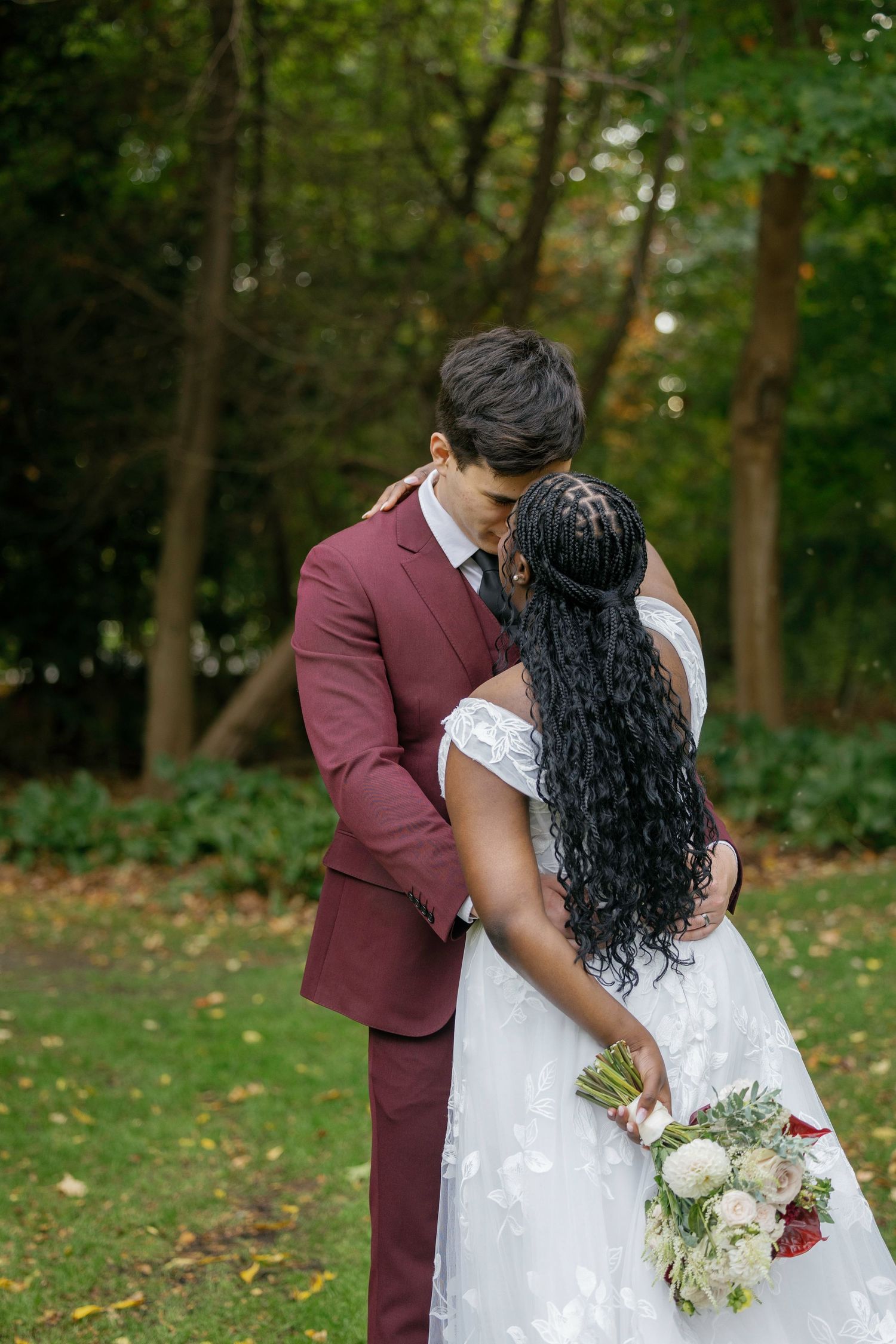 A bride and groom share a romantic kiss outdoors, she holds a bouquet while wearing a white gown and he wears a burgundy suit.
